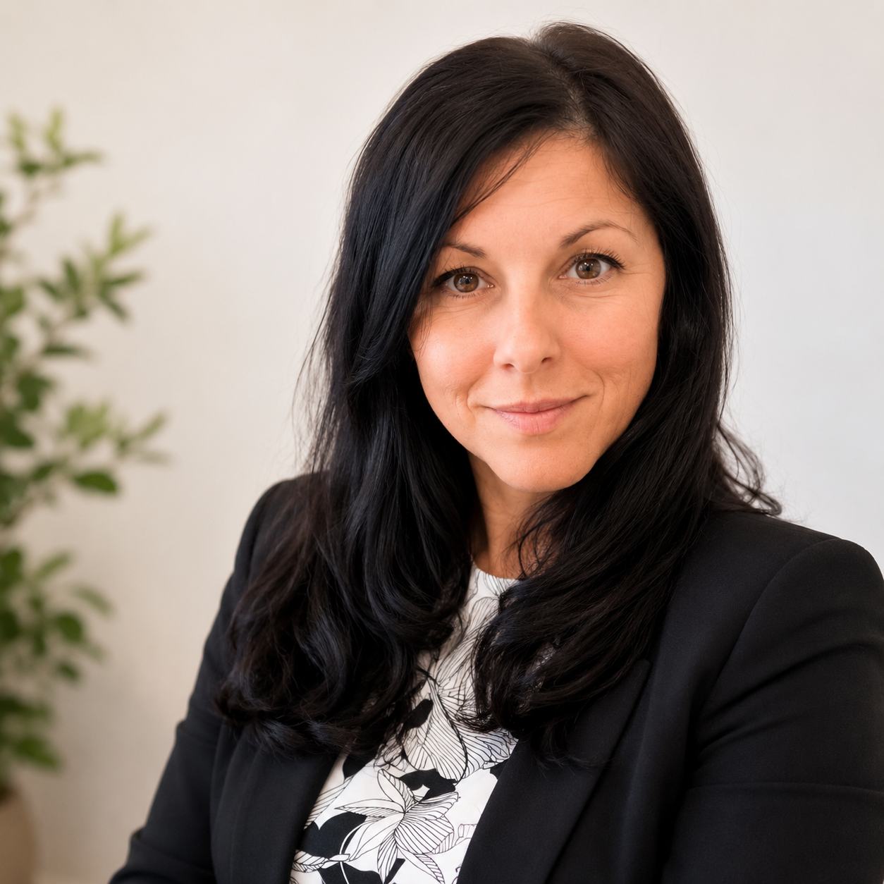 Portrait of a woman, Barbara Sok, with long black hair, wearing a black blazer and a floral blouse, smiling in front of a plain background with a plant on the side.