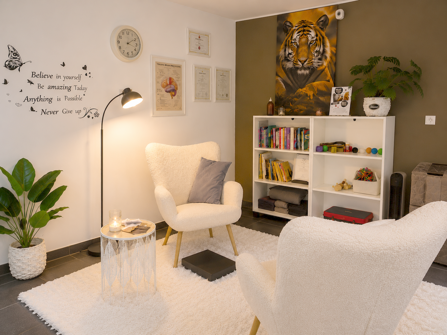 A cozy living room with two white armchairs, a white fluffy rug, and a small side table with a lit candle and a book. There is a tall black floor lamp next to one chair, a potted plant, a white bookshelf filled with books and small toys, and framed certificates and posters on the wall. A large tiger painting hangs behind the bookshelf, and a decorative wall quote reads: 'Believe in yourself, be amazing today, anything is possible, never give up.'