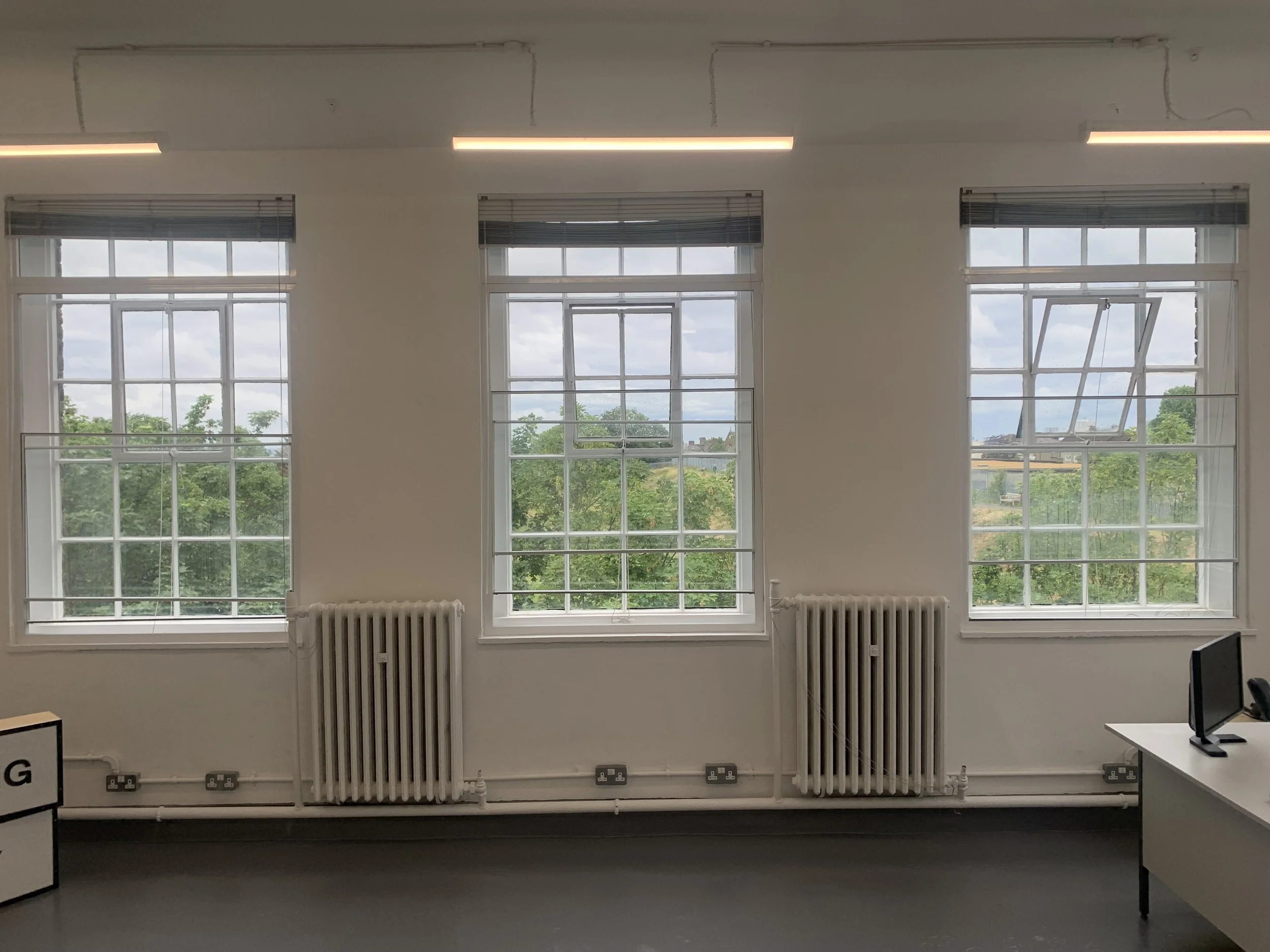 Interior of a room with three tall windows showing a cloudy sky and green trees outside. Two white radiators are positioned under the windows. On the left side, part of a sign with black text is visible, and to the right, a small section of a compute
