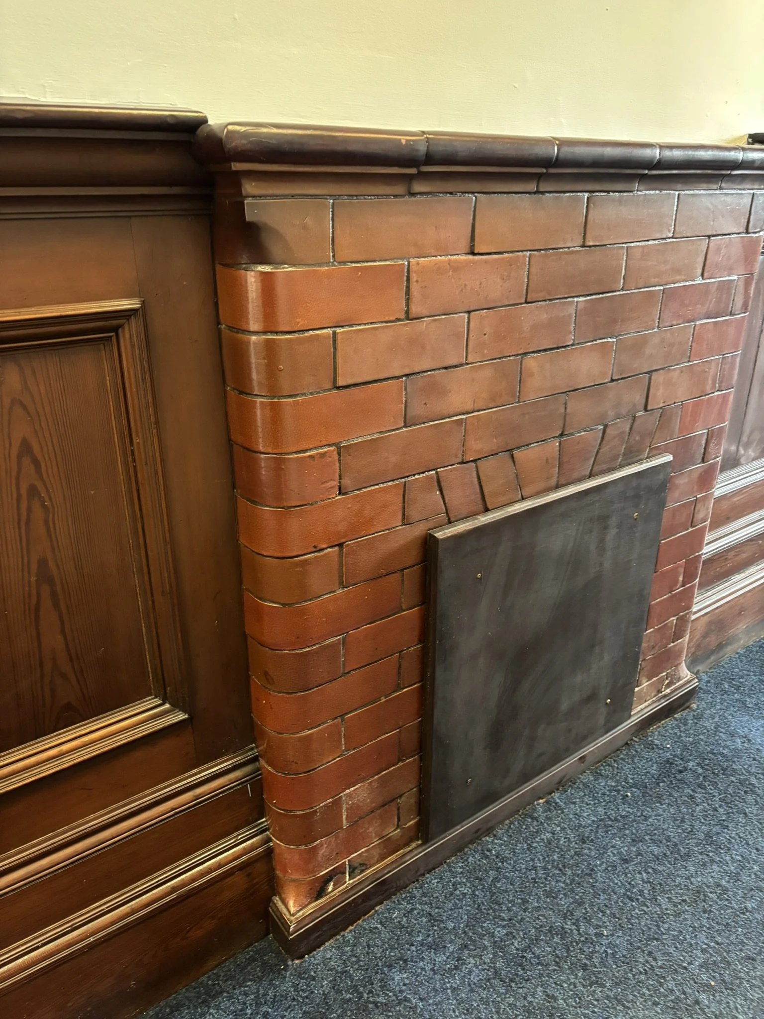 Close-up of a brick fireplace with a wooden mantle and paneling on the adjacent wall, with a dark baseboard and carpeted floor.
