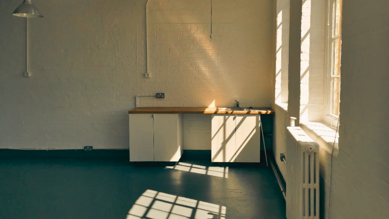 Sunlight streams through partially open window blinds, casting shadows on a white brick wall and a white kitchen cabinet with a wooden countertop.