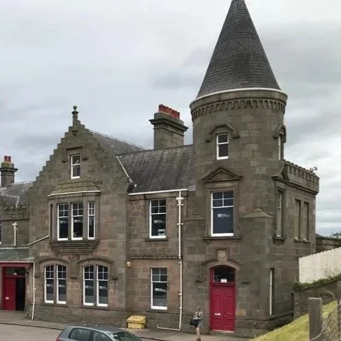 A historic stone building with a tower and turrets, featuring multiple windows, a red door, and a slate roof, under a cloudy sky.