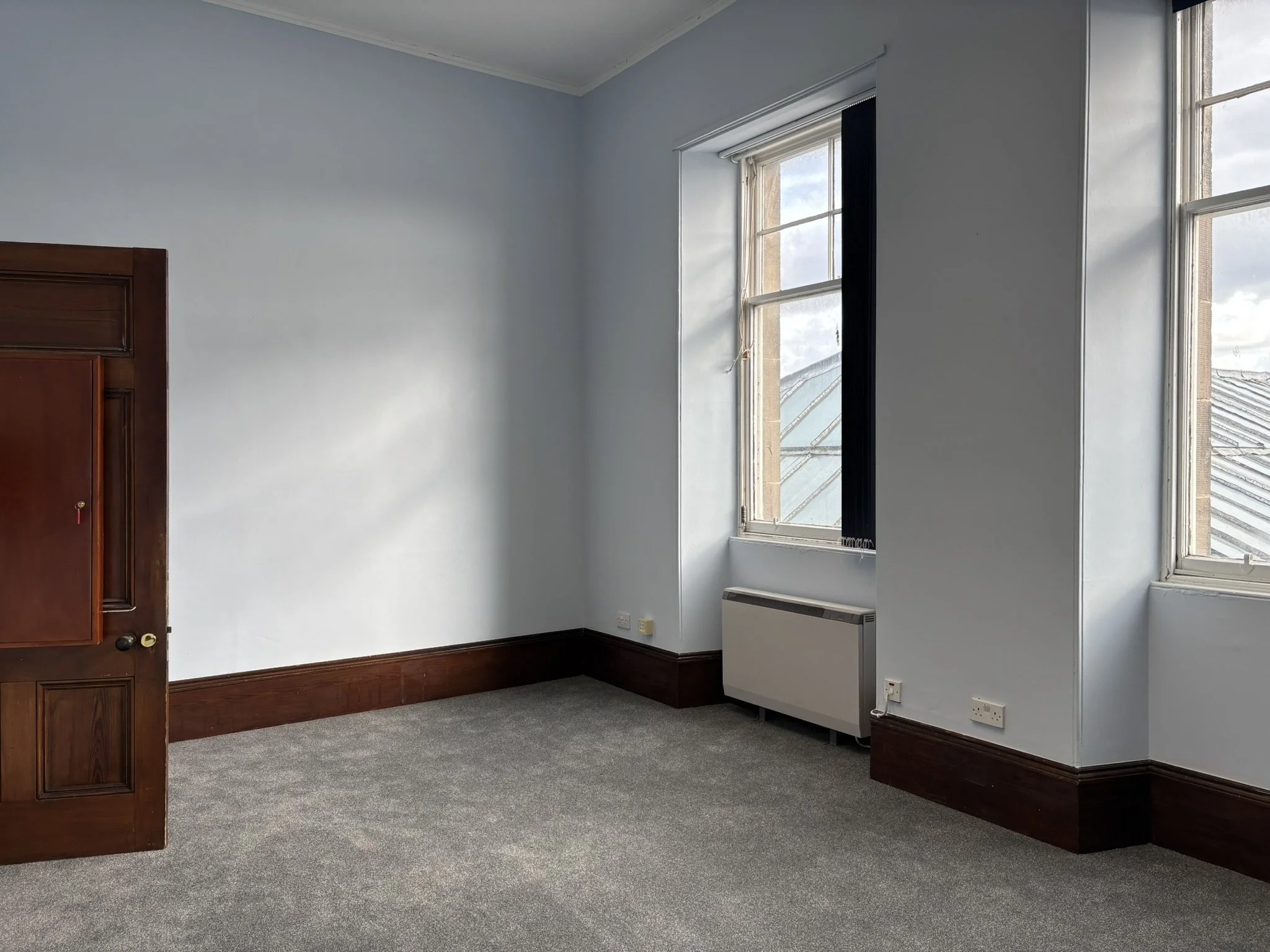 Empty room with light blue walls, dark borders, and a large window letting in natural light. There is a wooden door on the left and a white radiator below the window.