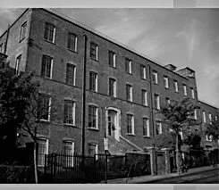 Black and white photo of a multi-story brick building with several windows, trees, and a fence in front.