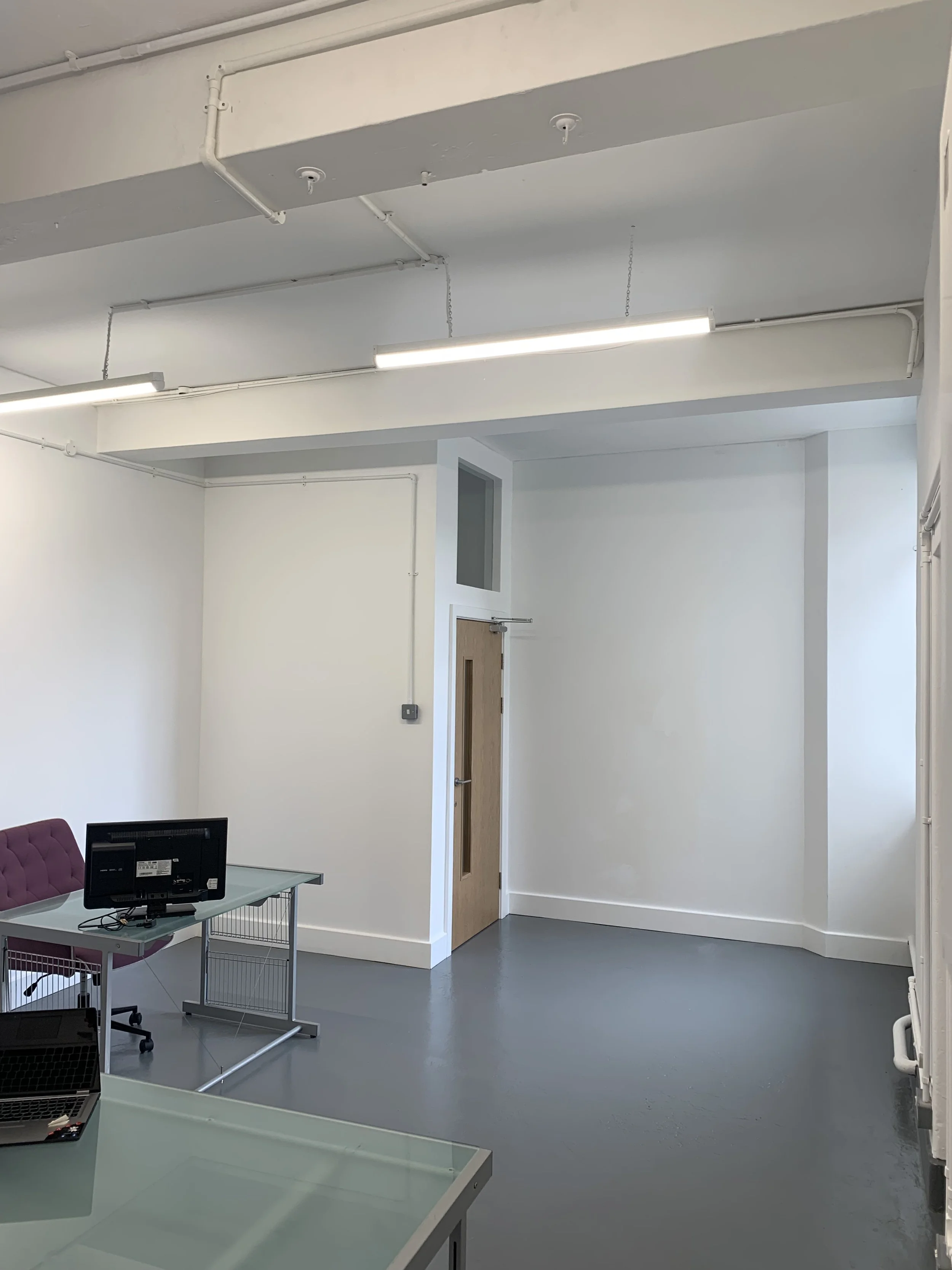 An office space with white walls, a wooden door, a small window, ceiling pipes, fluorescent lighting, and an office table with computers and a purple chair.