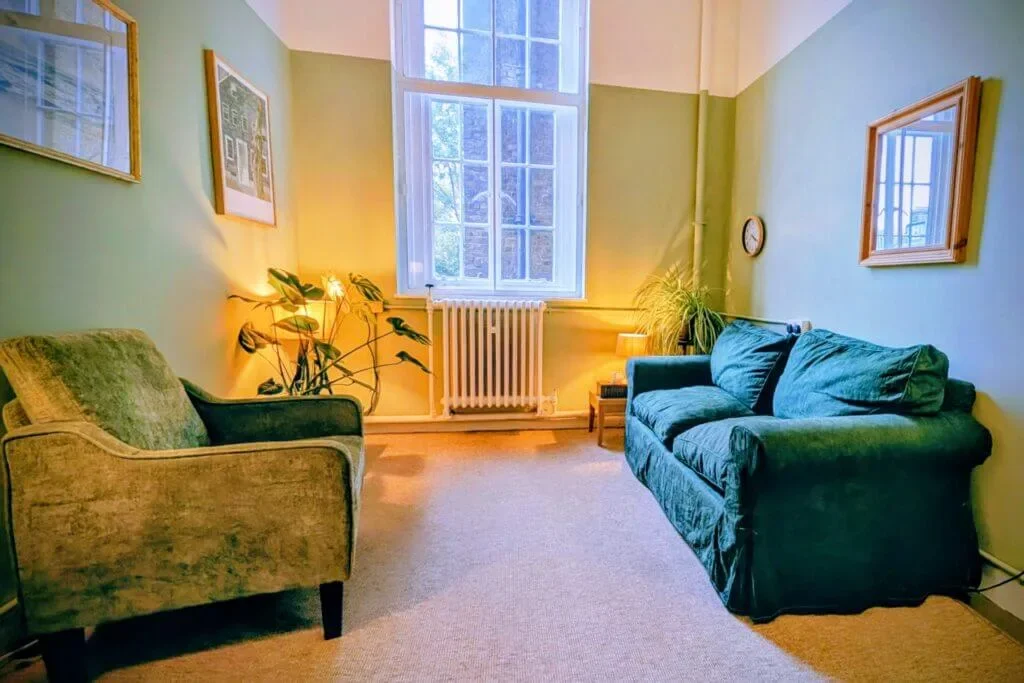 A living room with a green armchair, a blue couch, large window, framed pictures on the walls, and a radiator beneath the window. There are houseplants and lamps providing warm lighting.