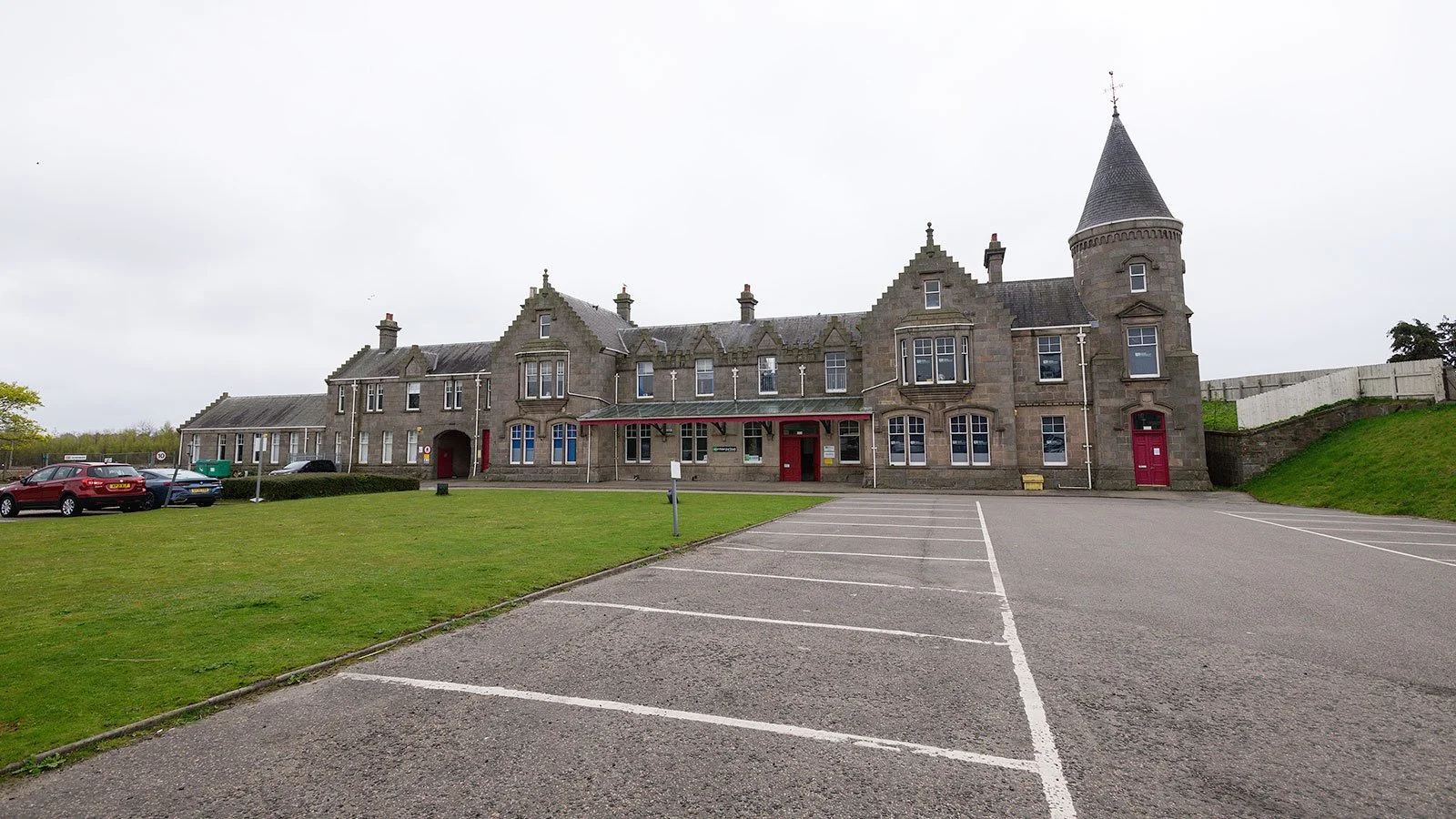 A large, historic stone building with a castle-like appearance, featuring a tower with a conical roof, multiple windows, and red doors, surrounding a parking lot and grassy area.