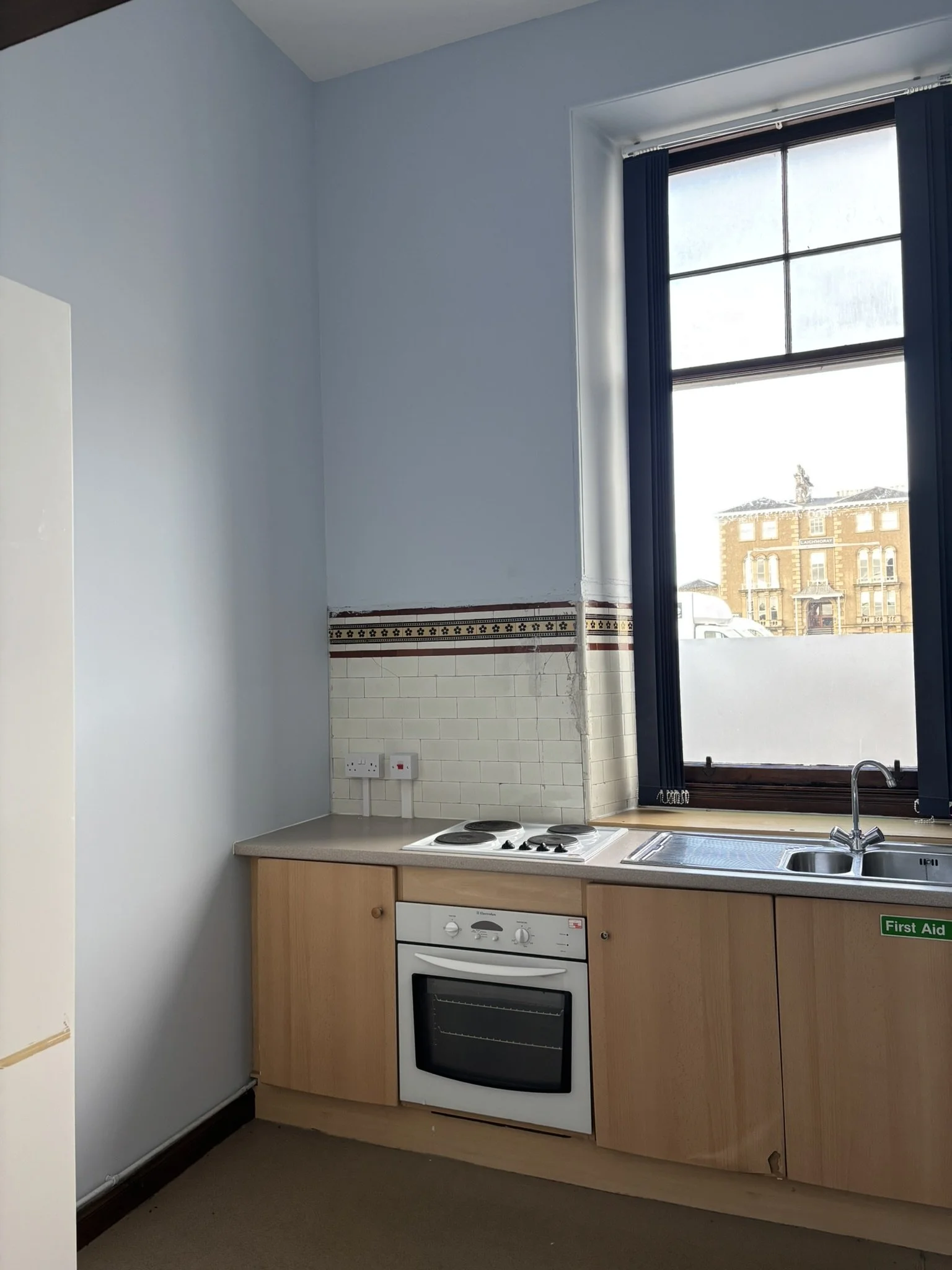 Kitchen with wooden cabinets, a built-in oven, a countertop with a four-burner stove, white tiled wall, a sink, and a window with blue curtains showing a building outside.