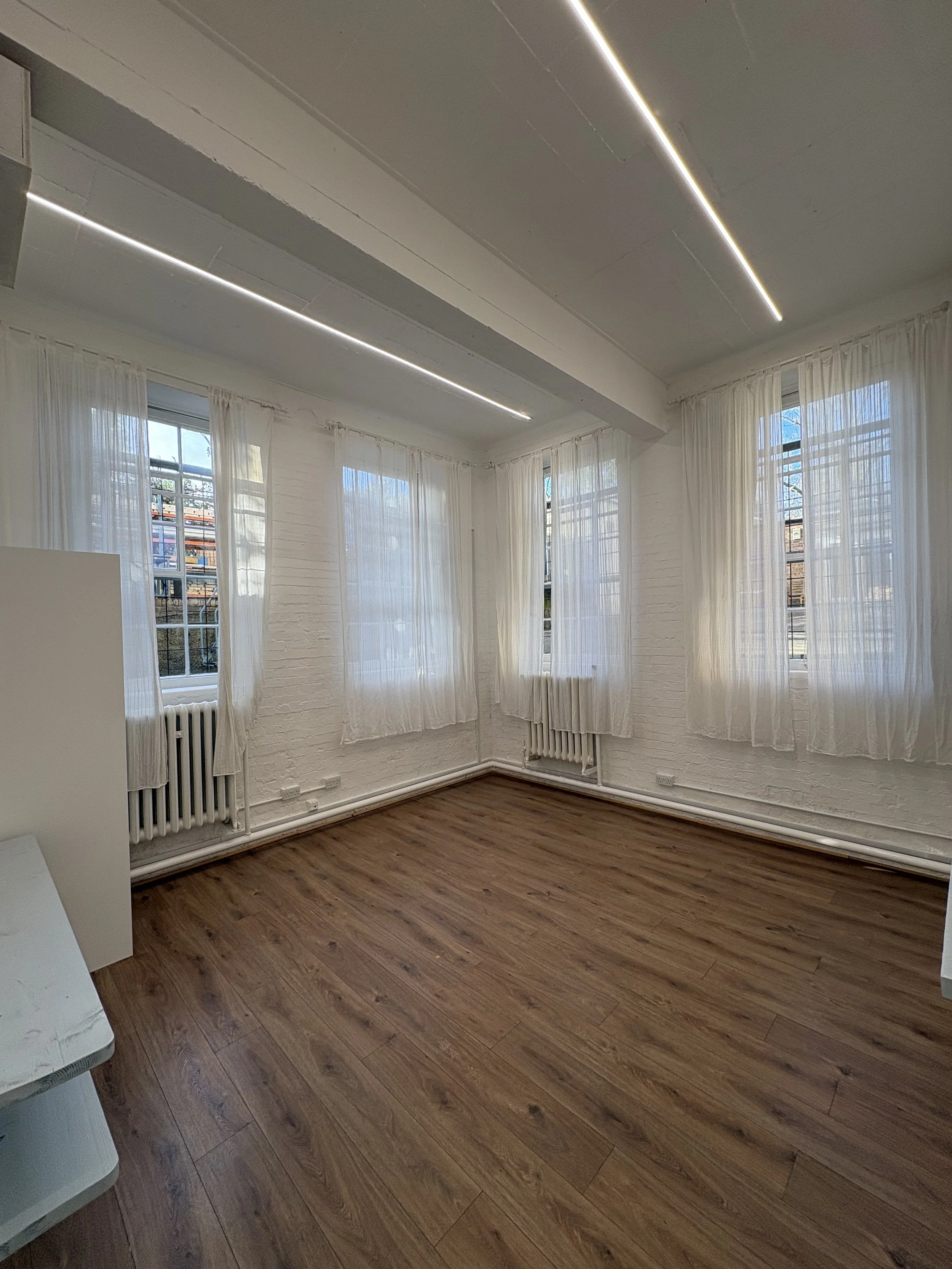 Empty studio with white painted brick walls, multiple large windows with sheer white curtains, wood floors, and ceiling with linear LED lighting.