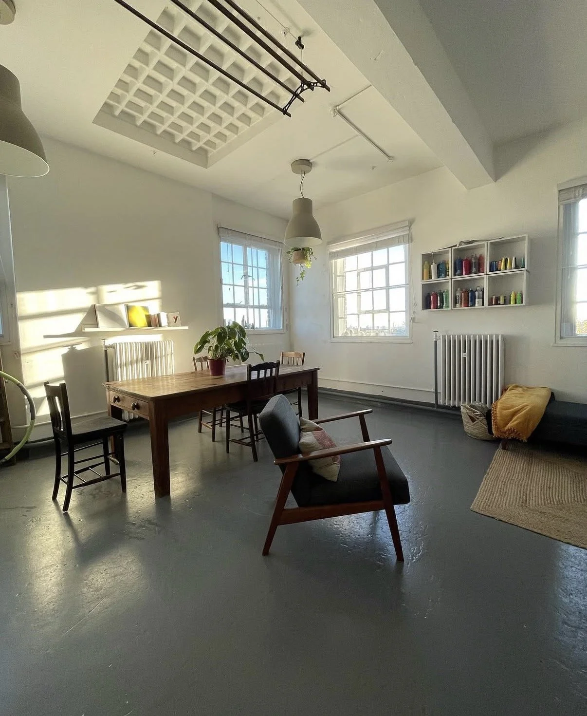 A cozy living room with natural light, a wooden dining table with chairs, a black armchair with a cushion, a yellow blanket on a gray couch, a small bookshelf with colored books, a potted plant, and large windows letting in sunlight.