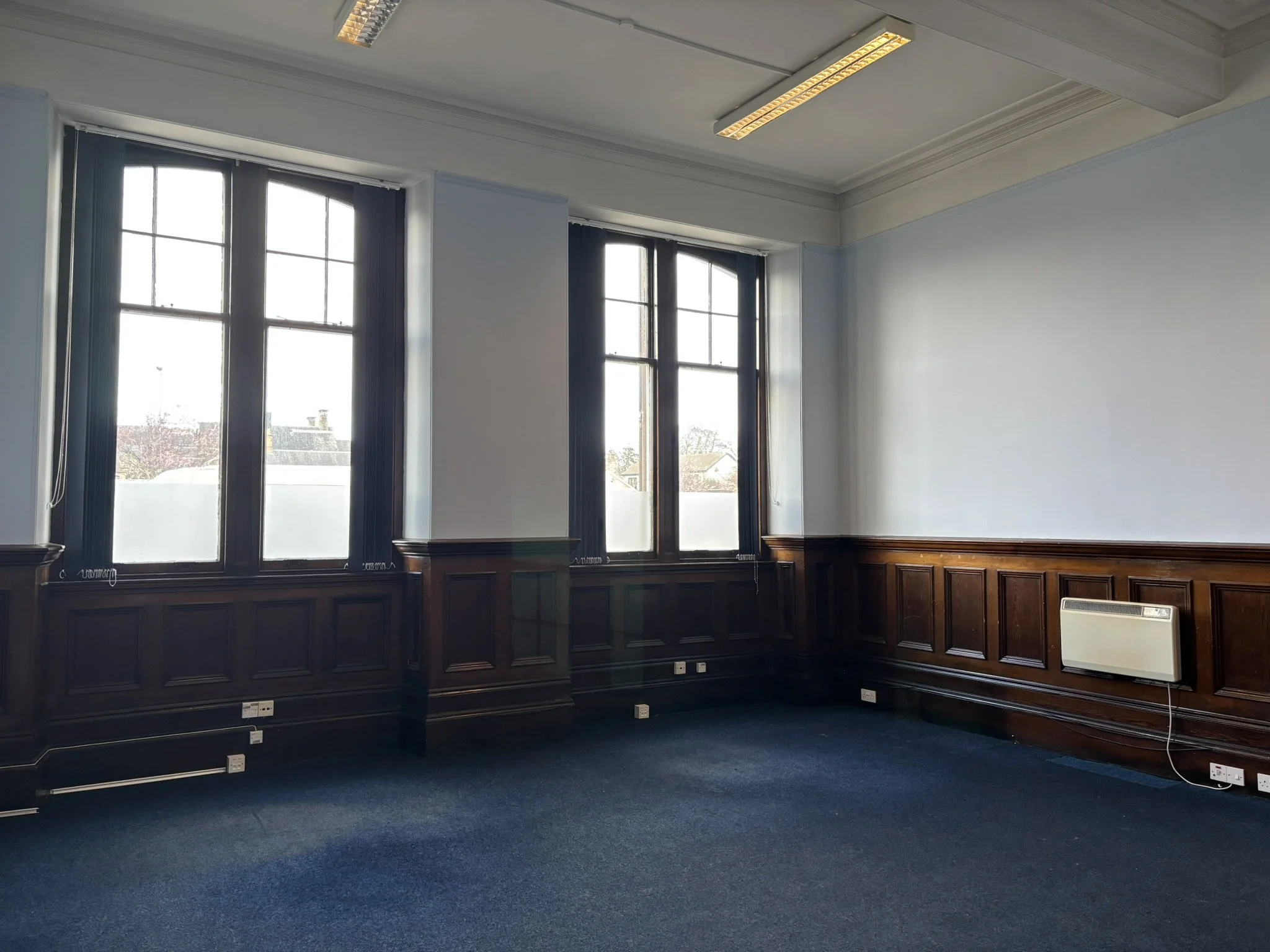 Empty room with large windows and dark wood paneling, blue carpet, and electrical outlets.