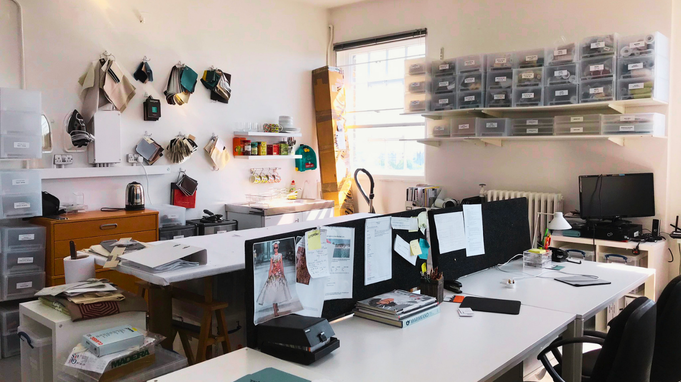 office space with white desks, a computer, and various office supplies. Behind the desks, there are transparent storage boxes on shelves, a window with curtains, and a wall with hanging fabric samples. There are also notebooks, papers, and sticky notes on the desks.