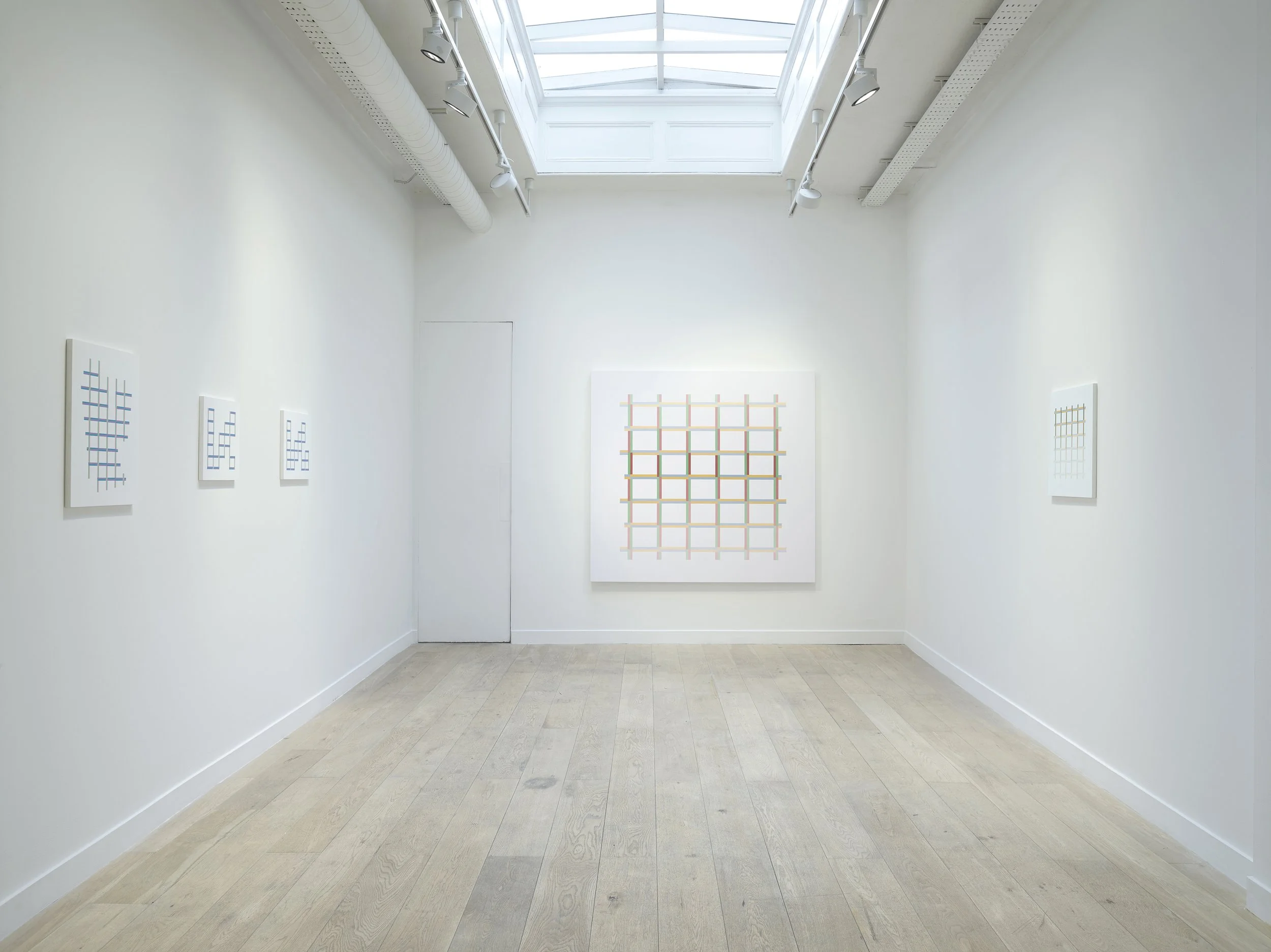 An art gallery with white walls, a light wood floor, and artworks displayed on the walls, illuminated by natural light from a large skylight ceiling.