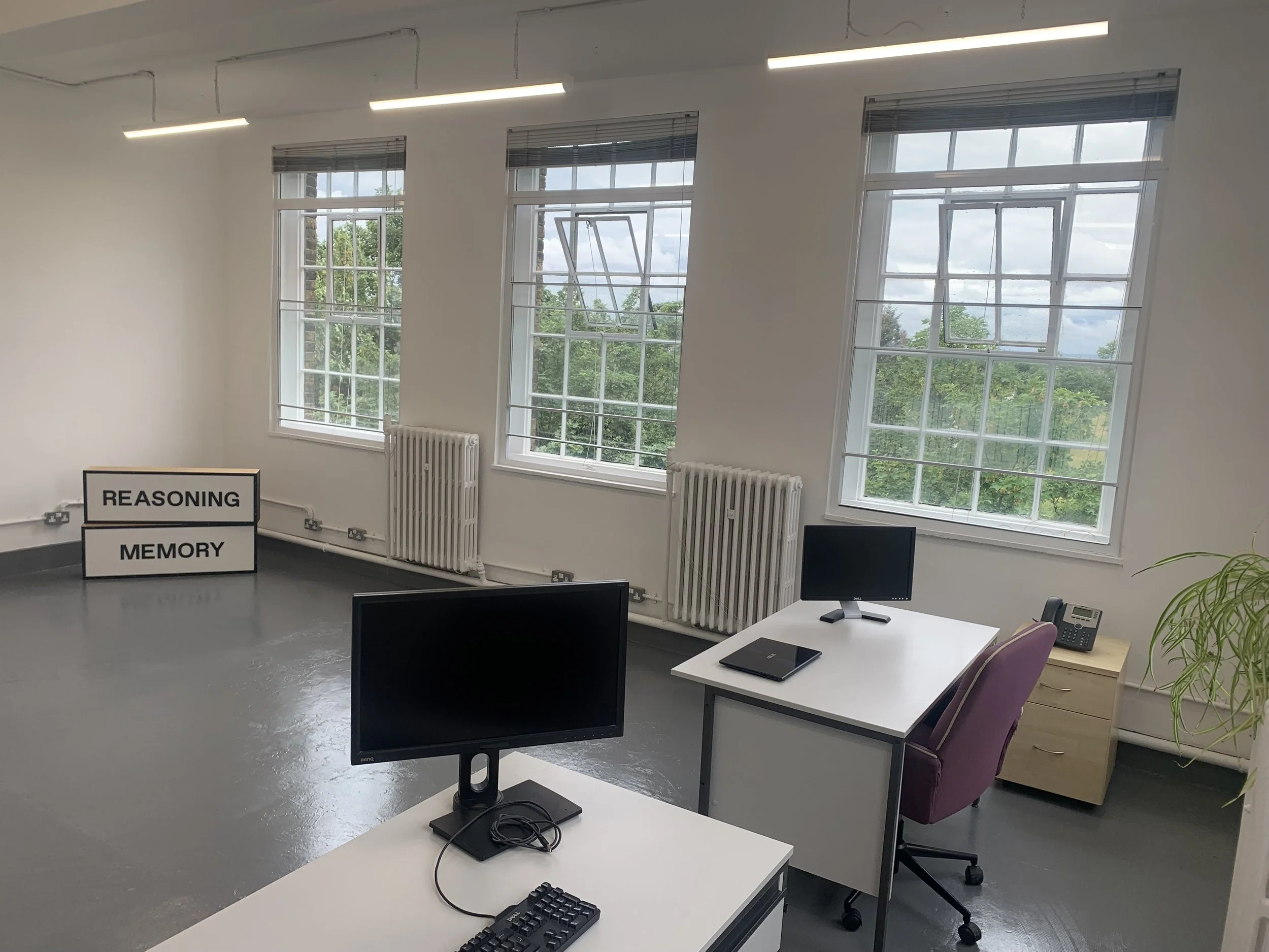 An empty office space with several windows, desks with computer monitors, a keyboard, a notepad, a desk phone, purple chairs, and a sign that reads "Reasoning" and "Memory".
