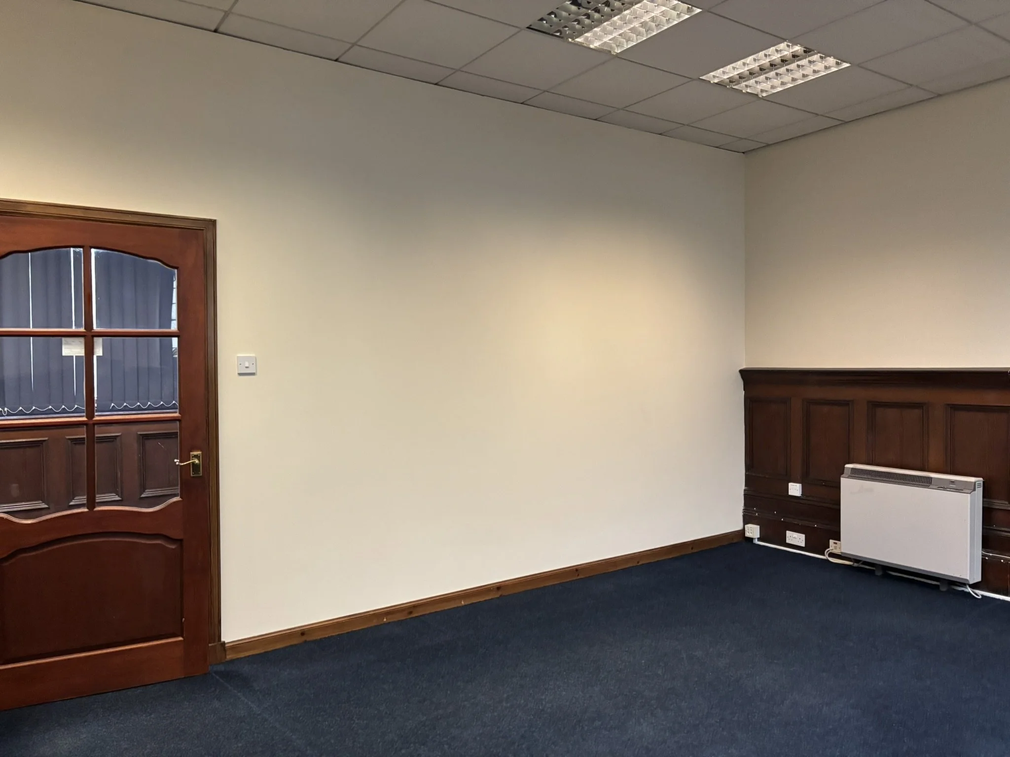 Empty room with a wooden door, beige walls, dark blue carpet, and a wall heater along with multiple electrical outlets.