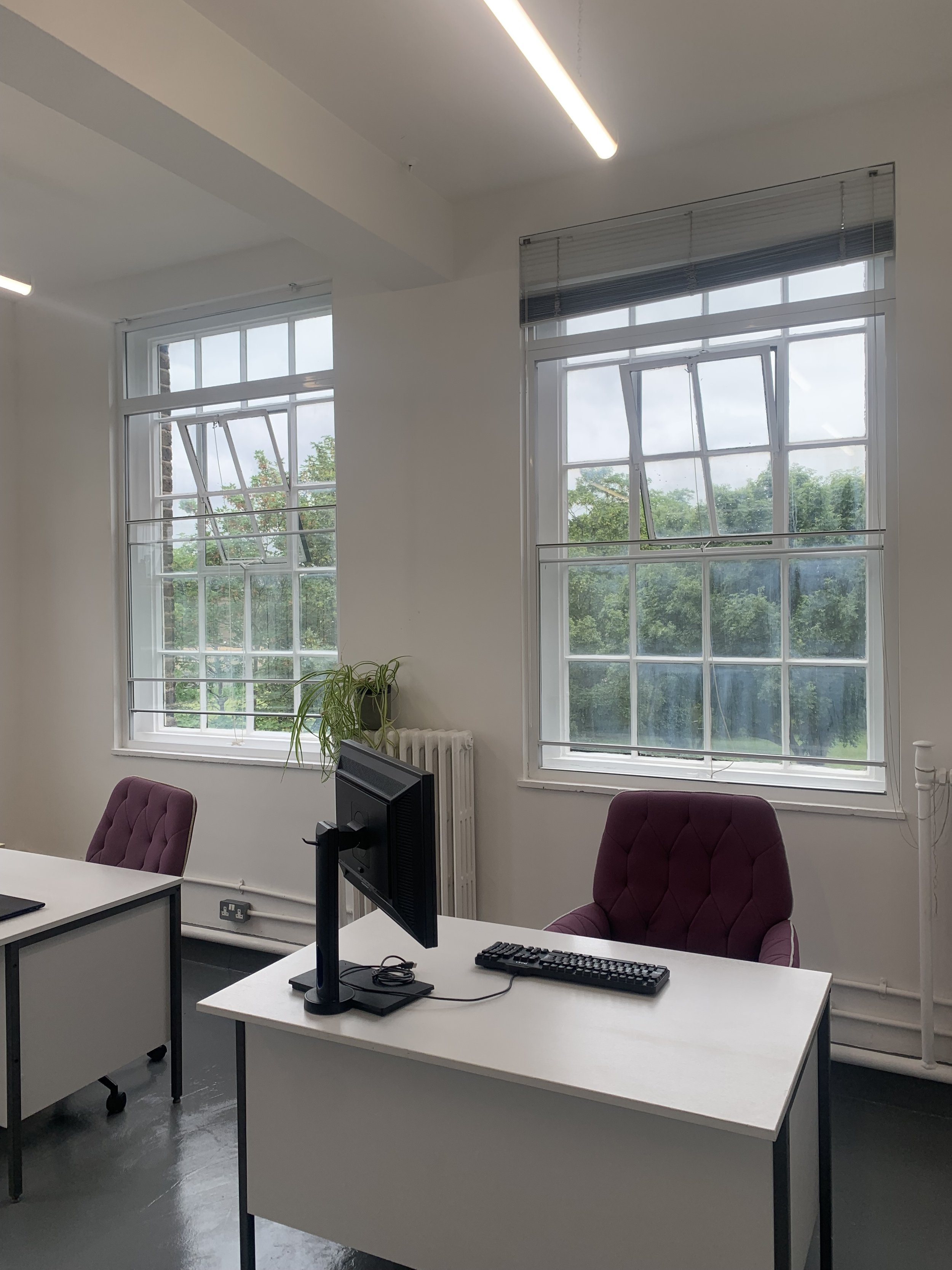 An office with two white desks and a purple cushioned chair, computer monitor, keyboard, and mouse. Large windows with blinds, a plant, and a radiator are visible in the background.