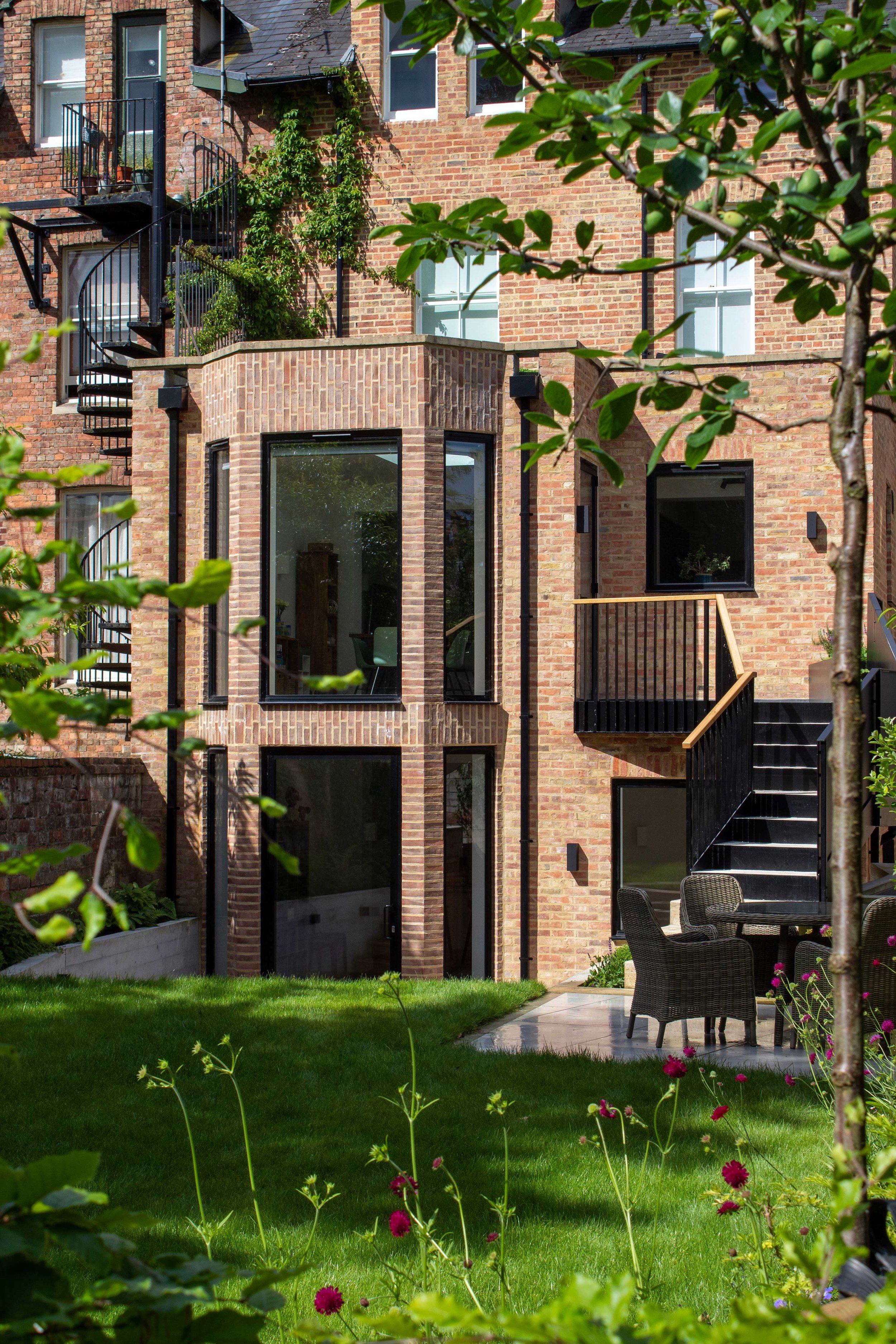 A brick apartment building with large windows and a black metal staircase on the left side. The building features balconies with black railings. In the foreground, there is a lush green lawn with various plants and flowering flowers. On the right side, there is an outdoor patio area with outdoor chairs and a small table.