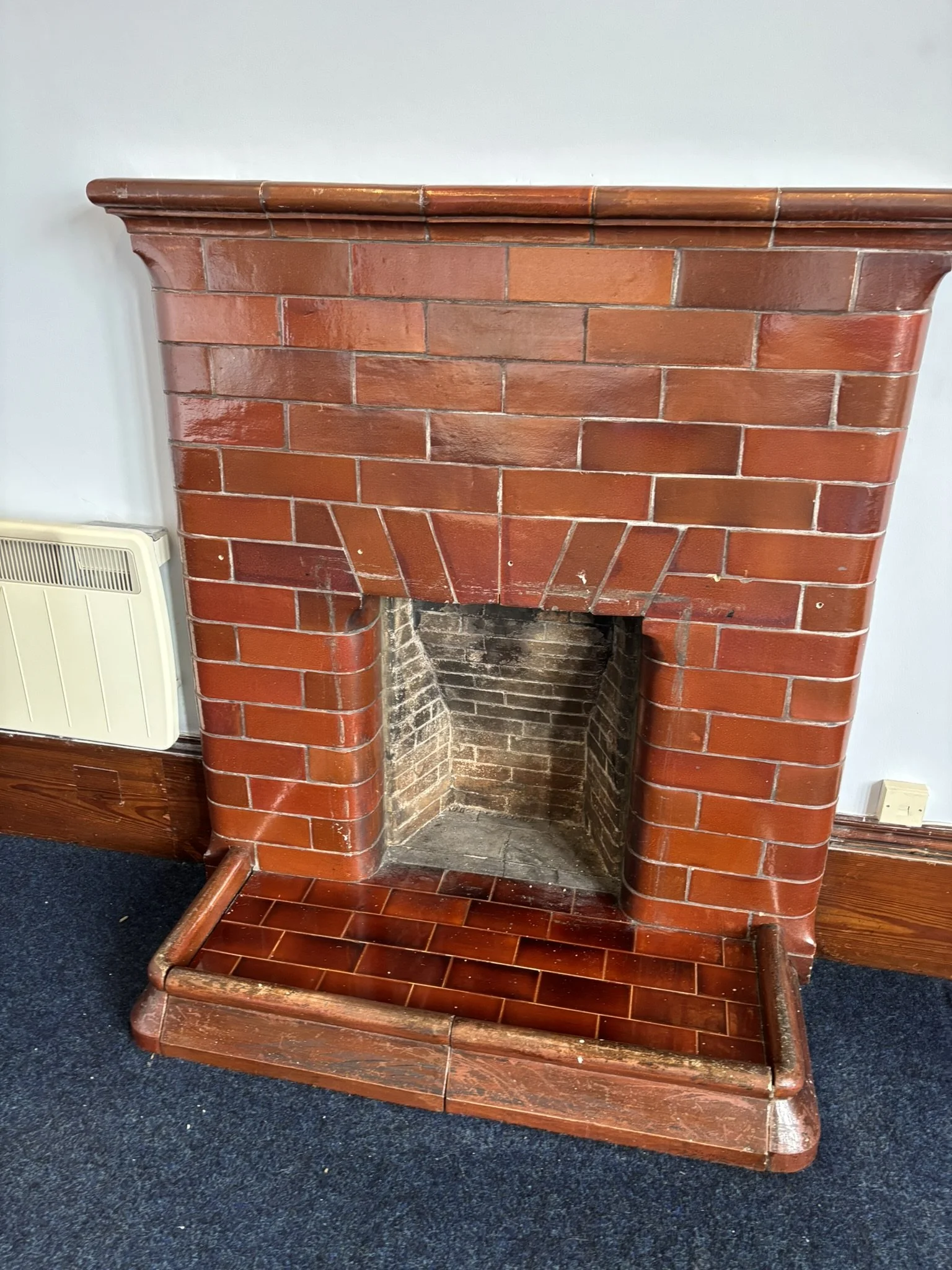 Empty red brick fireplace with a wooden mantel and a metal frame at the base, situated on a wooden floor with a dark carpet nearby.