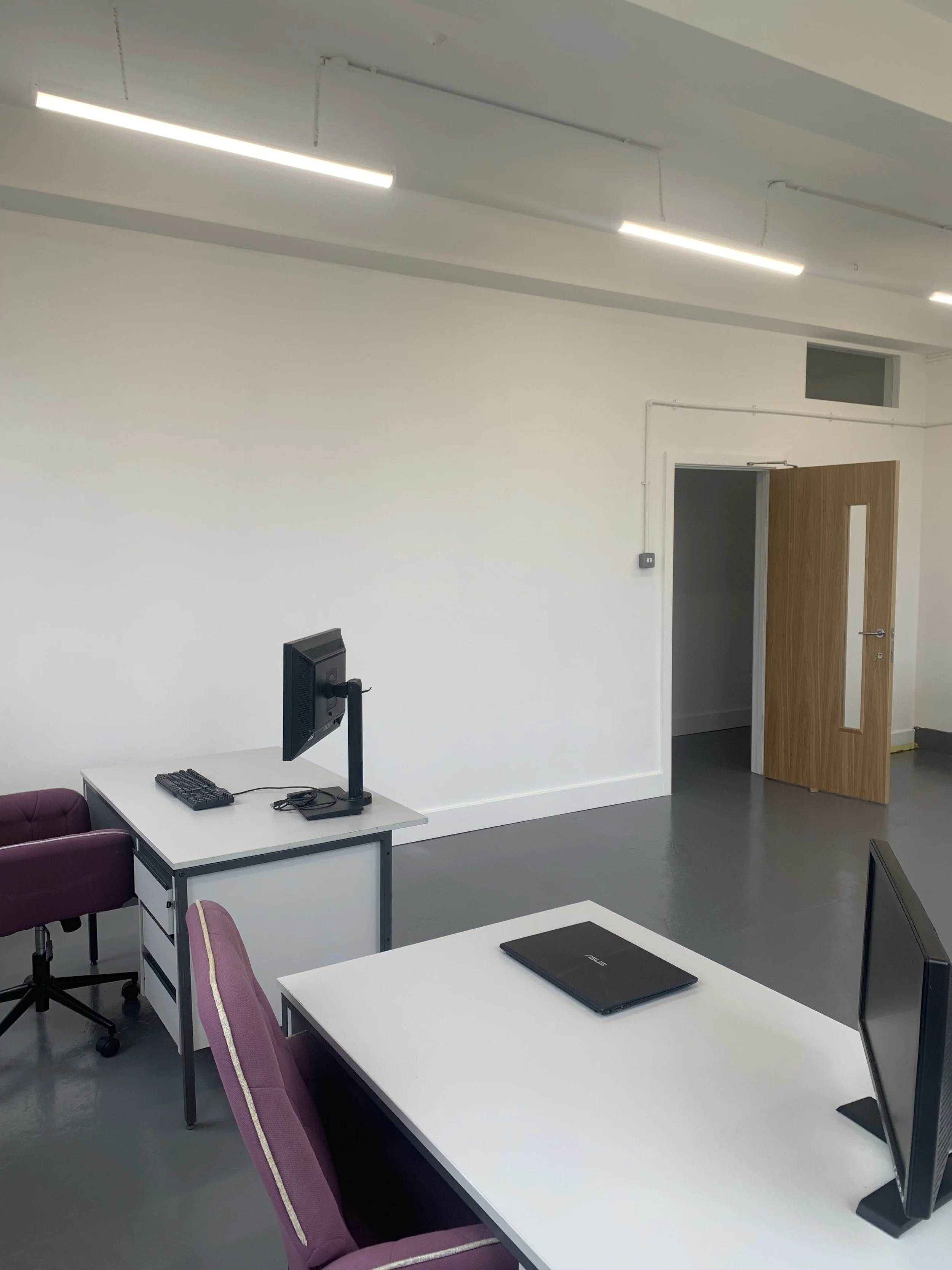 An empty classroom or office with white walls and ceiling, fluorescent lights, several white desks with purple chairs, desktop computers, a closed black laptop, and an open wooden door.
