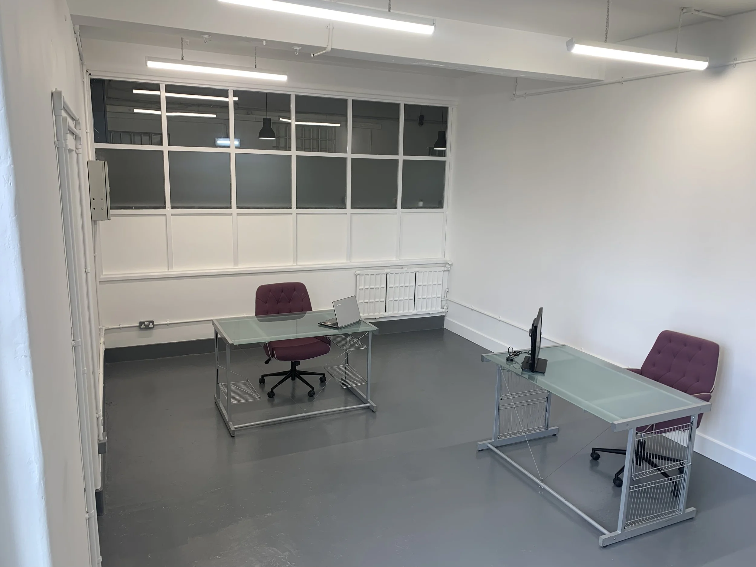 An empty office or conference room with white walls, gray flooring, and a large windowed partition. The room contains purple chairs, glass desks, a laptop, a computer monitor, and wire storage carts. Ceiling lights and a radiator are also visible.