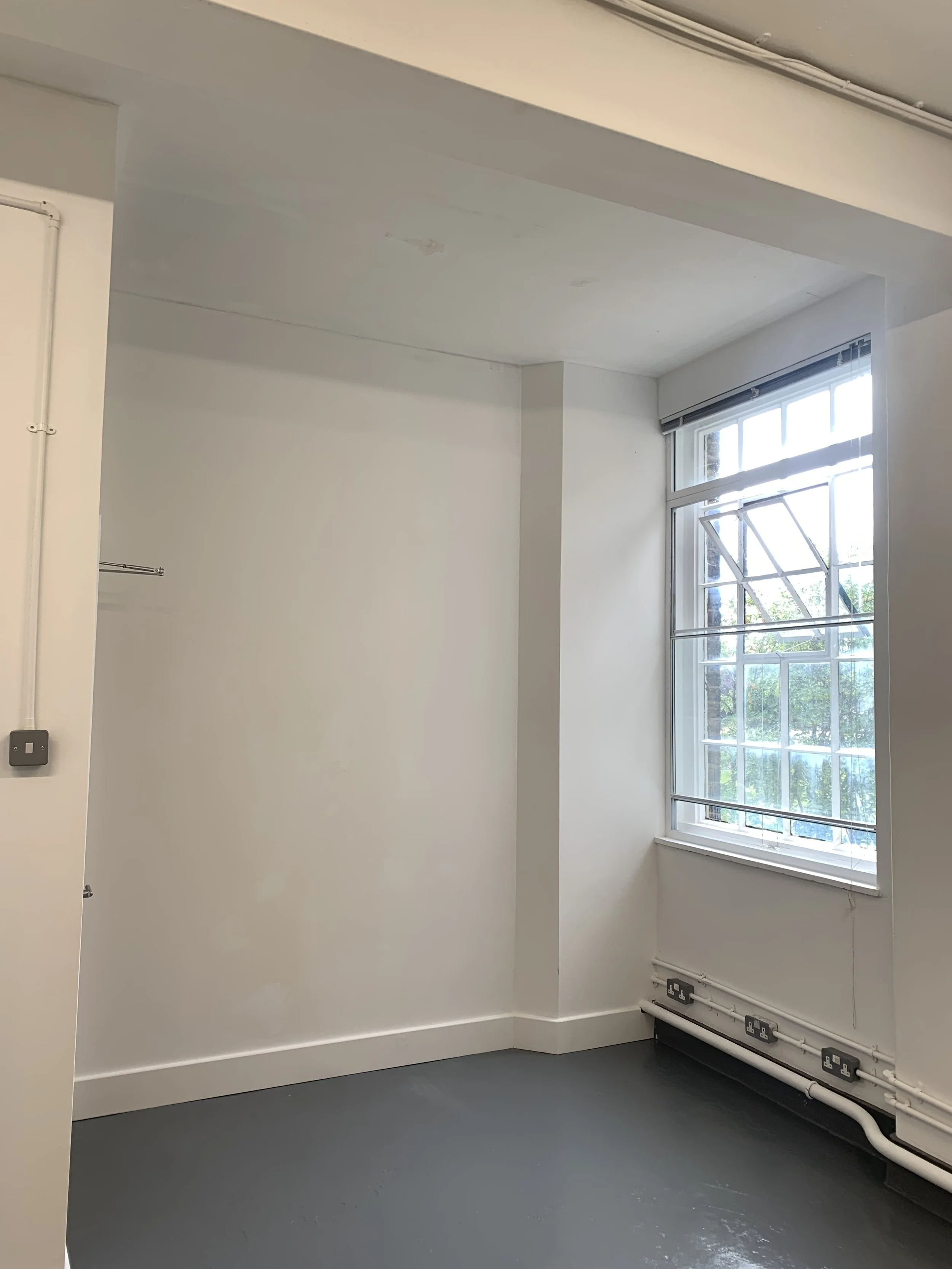 A clean, minimalistic room with white walls, a large window with partially open blinds allowing natural light, and electrical outlets along the bottom of the wall.