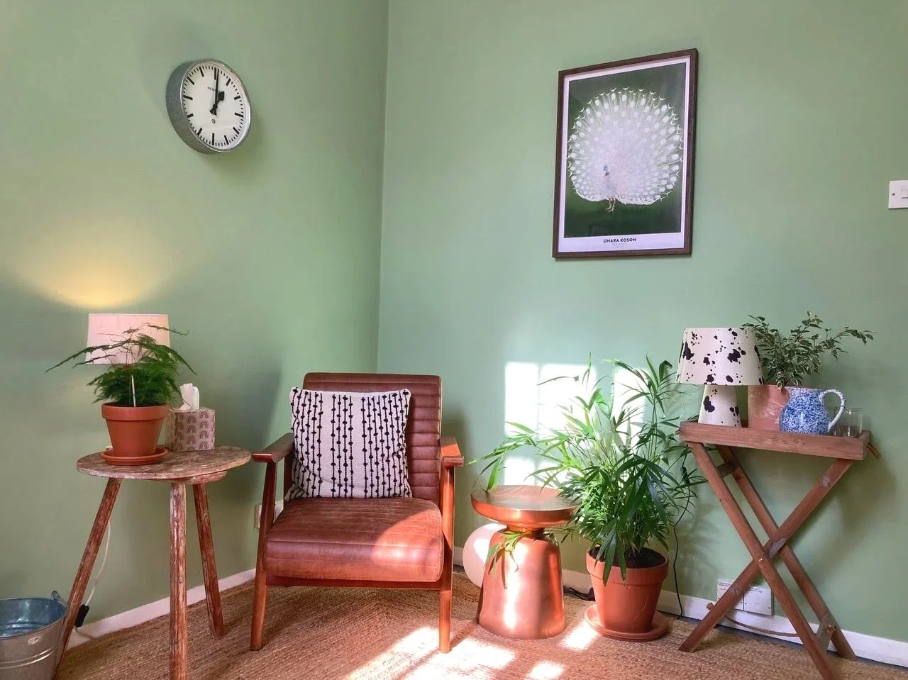 Living room corner with a green wall, clock, framed peacock poster, wooden chair with a patterned cushion, small wooden side table, potted plants, and a table lamp with a flower pattern shade.