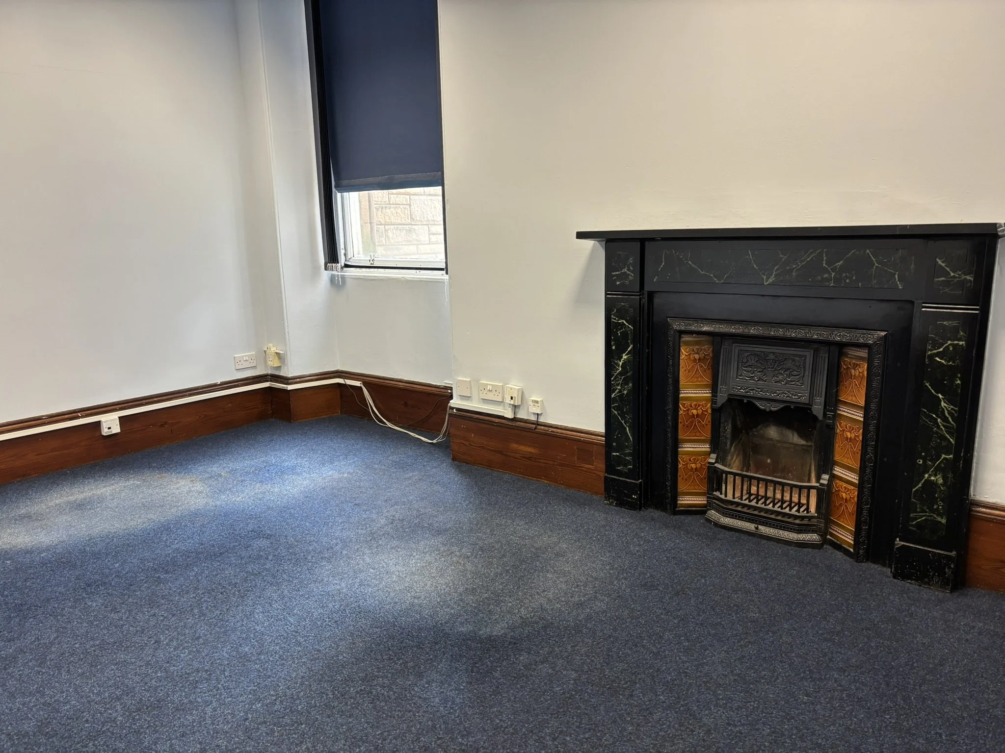 Empty room with blue carpet, white walls, black fireplace with decorative orange tiles, and a window with a blue roller blind.