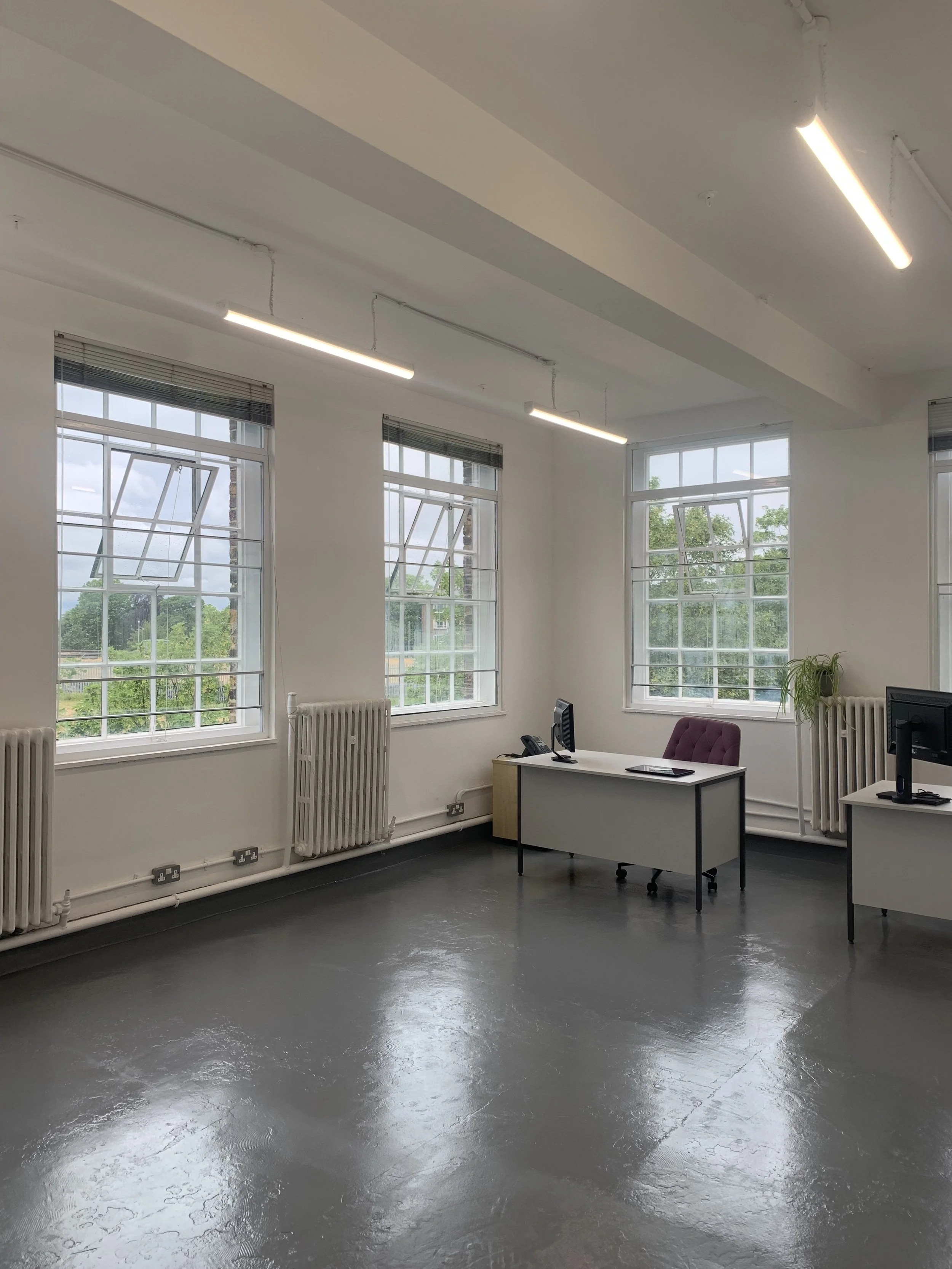 An empty office with white walls, large windows with blinds, and fluorescent lights on the ceiling. There are radiators beneath the windows, a desk with a computer, phone, and chair, and a potted plant in the corner.