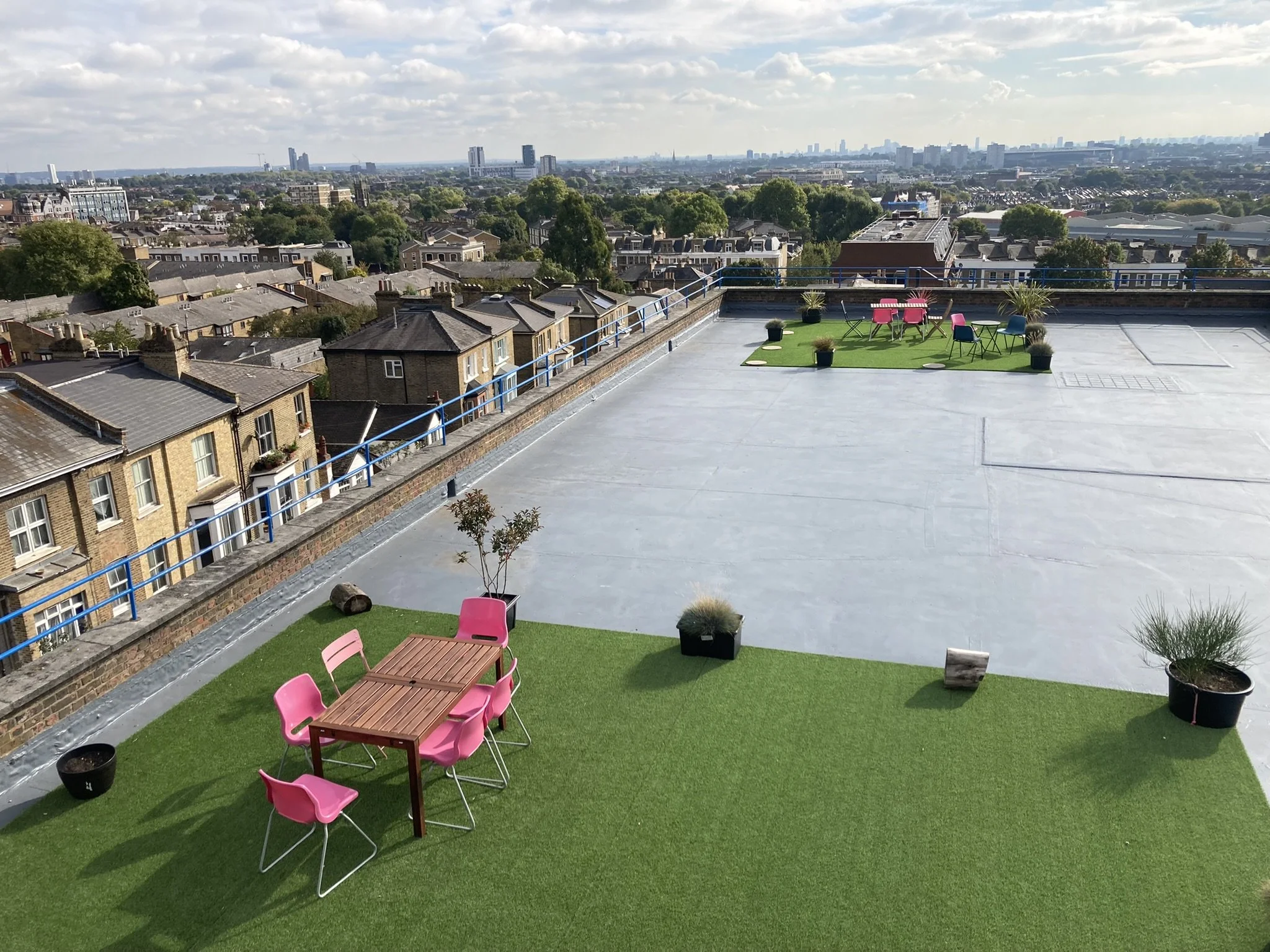 Rooftop terrace with artificial grass lawn, pink and blue chairs, wooden table, potted plants, and cityscape view with buildings and trees under partly cloudy sky.