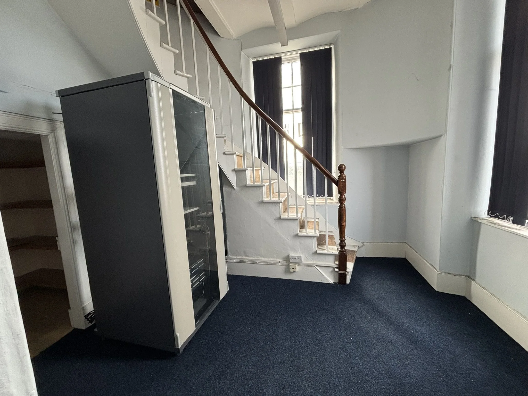 Interior of a house with carpeted floor, staircase with wooden handrail, and a black vending machine or refrigerator near a doorway. There is a large window with black blinds letting in natural light.