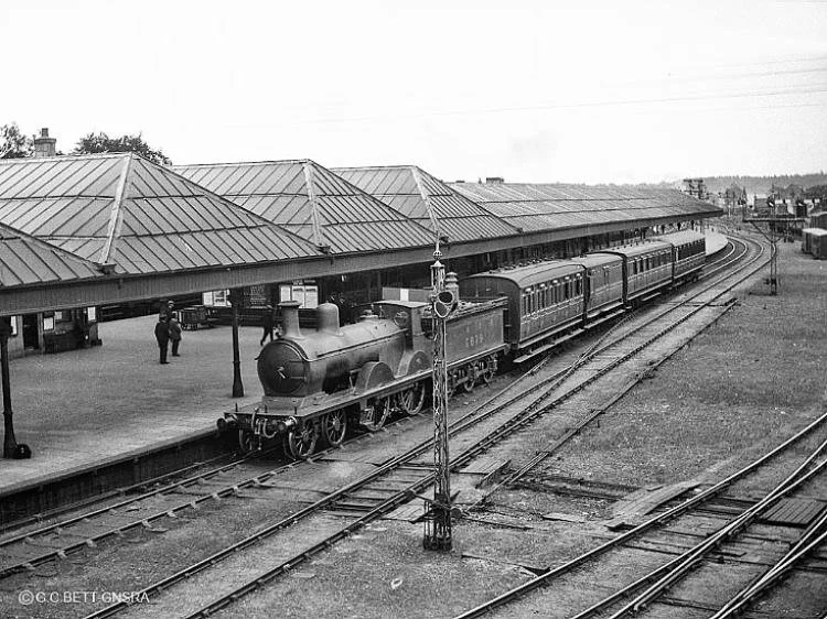 Vintage black-and-white photo of a steam train arriving or departing a train station with a covered platform, passengers waiting, and a rural landscape in the background.