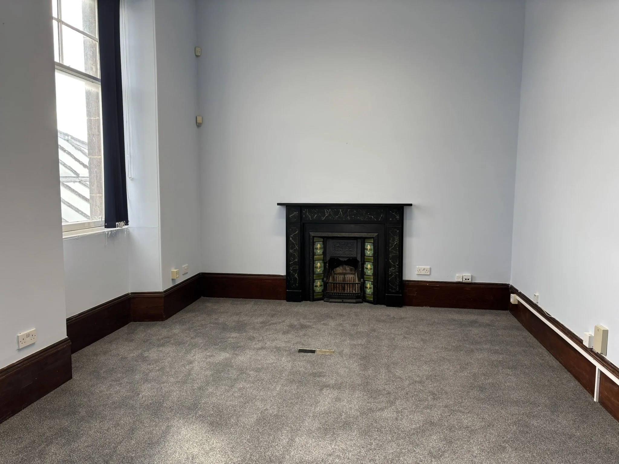 Empty room with gray carpet, white walls, a black fireplace with decorative tiles, a large window with vertical blinds, and electrical outlets along the baseboards.