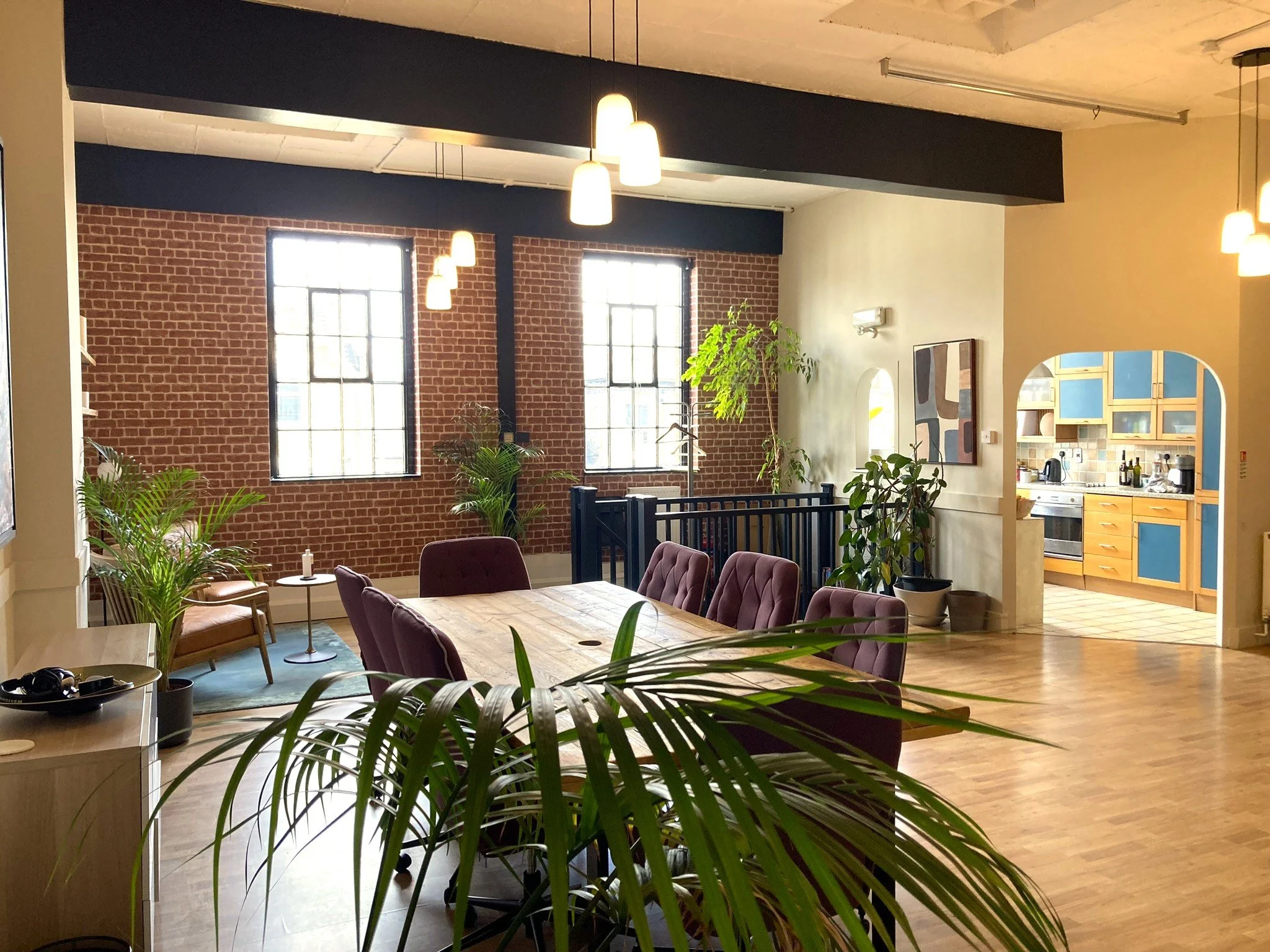 Interior of a modern apartment or office space with brick walls, large windows, hanging pendant lights, a wooden dining table, purple chairs, green indoor plants, and a view into a kitchen area with wooden cabinets and a tiled backsplash.