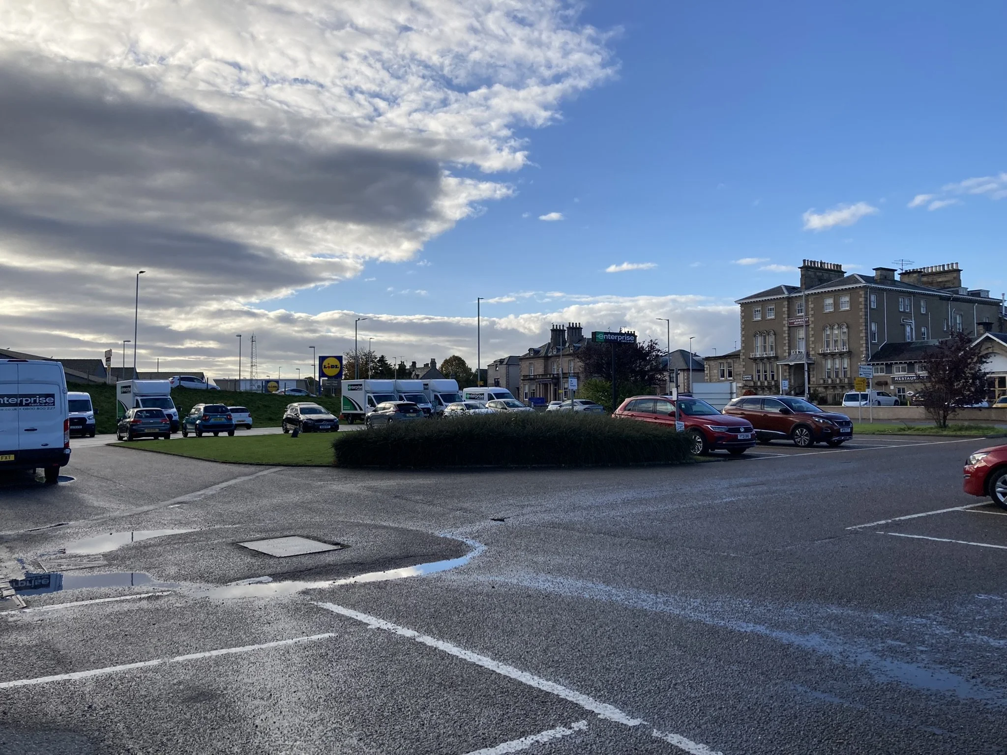 A parking lot with cars and vans, some puddles on the ground, behind which is a street with houses, a supermarket sign, and a partly cloudy sky.
