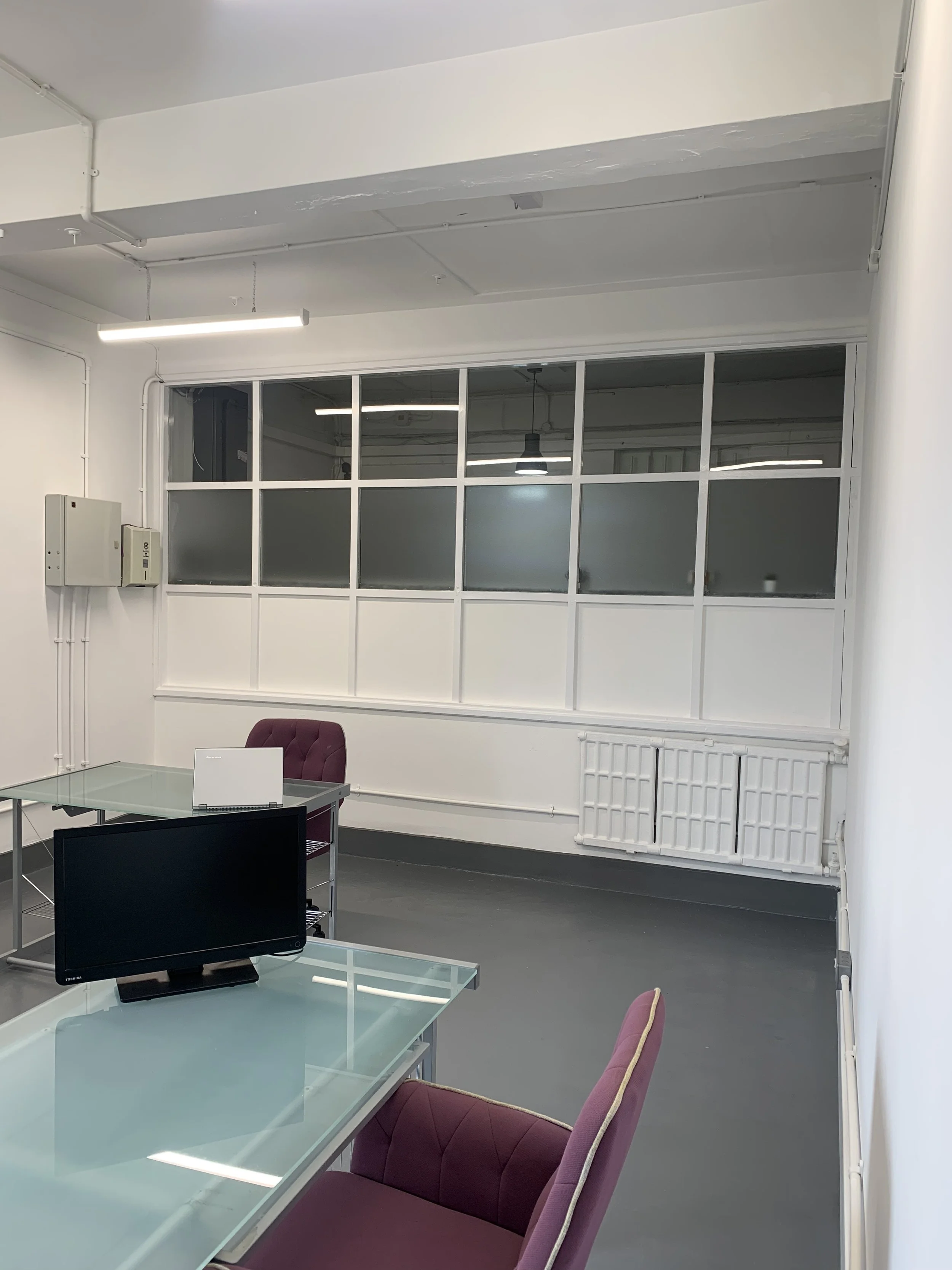 An office space with a large glass desk, pink chair, computer monitor, and windowed partition wall.