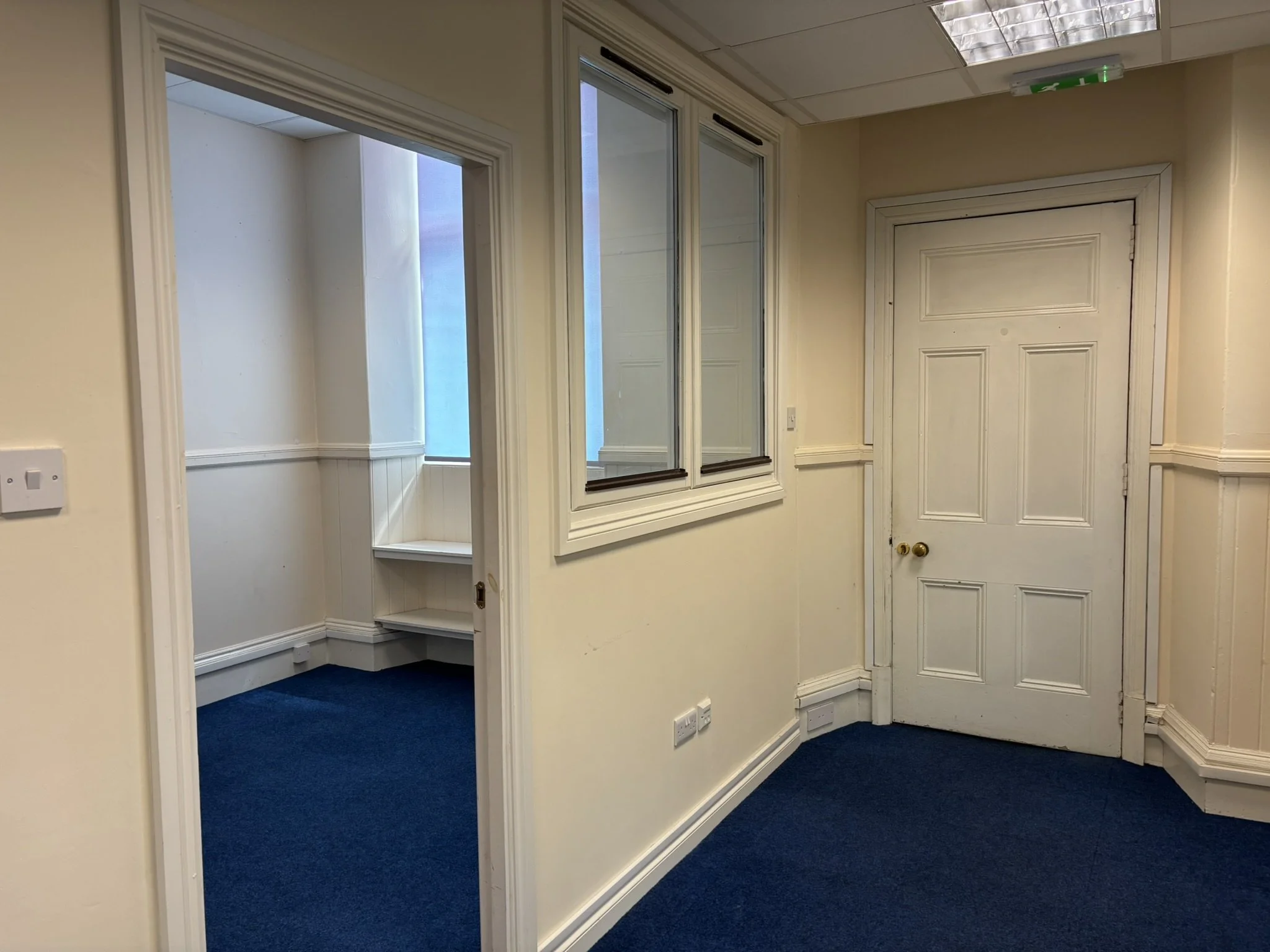 Interior of an office space with a white door, a wall-mounted mirror, cream-colored walls, and blue carpeted floor.
