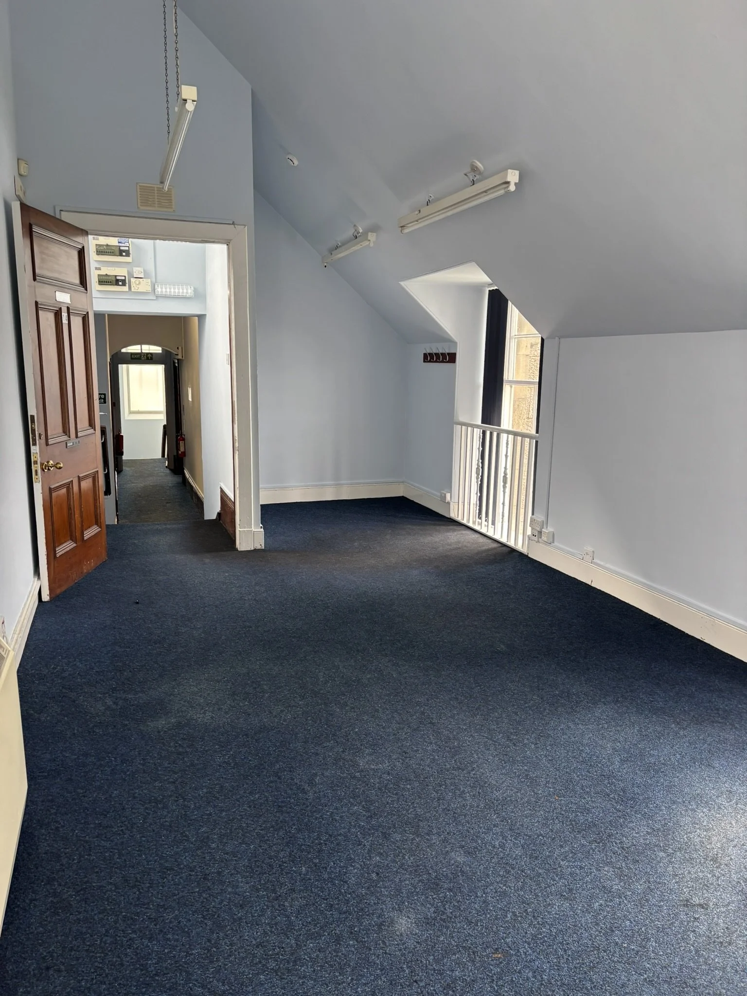 Empty room with blue carpet, white walls, small window, and open wooden door leading to a corridor.