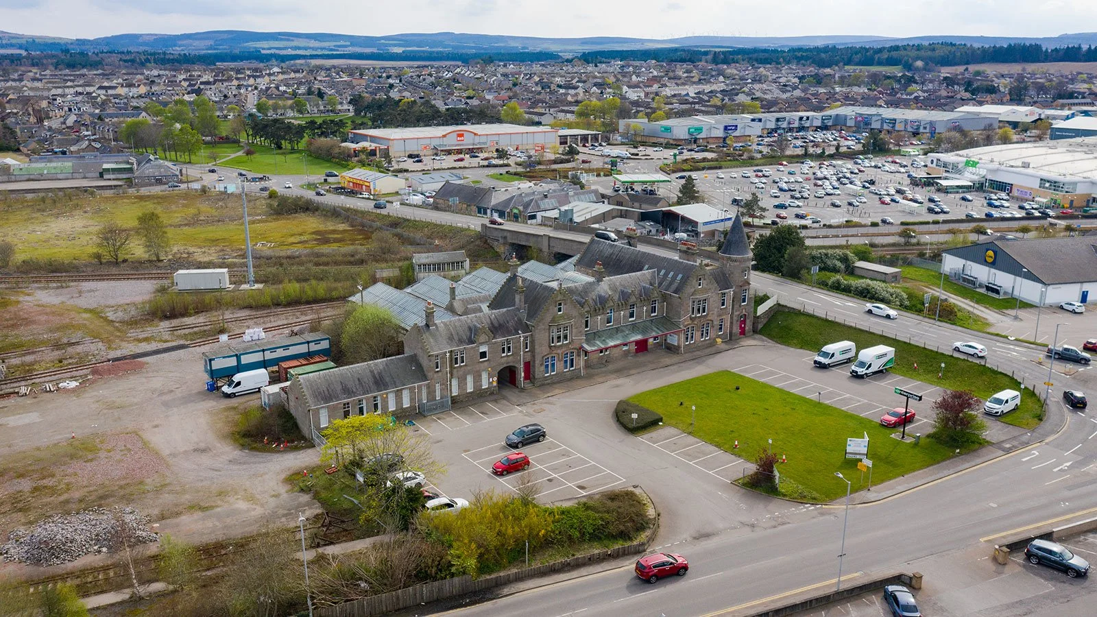 Aerial view of a historic stone building with a castle-like turret, surrounded by parking lots, open green spaces, and a large shopping center in the background. The scene includes cars, trucks, and trees.