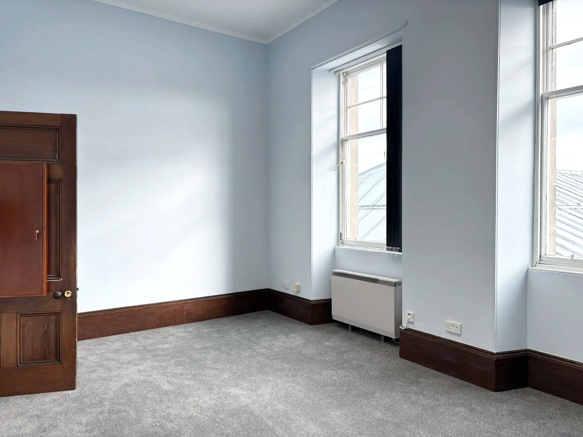 An empty room with light blue walls, a large window with white trim, a gray carpeted floor, a wooden door, and a white radiator near the window.