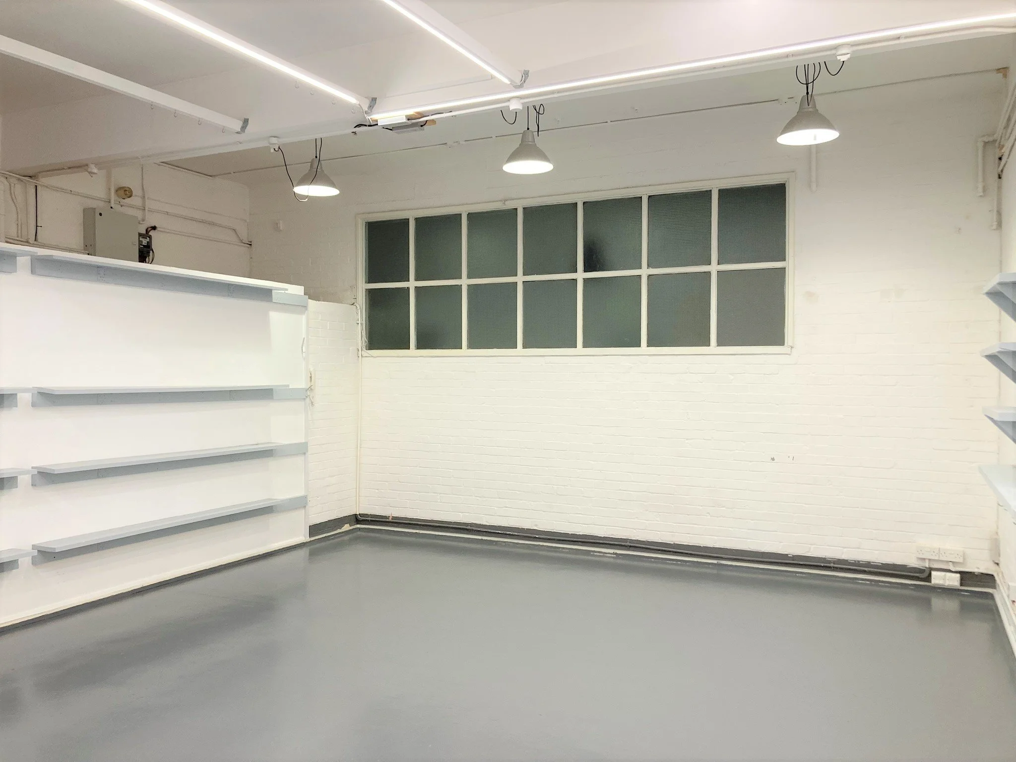 Empty retail space with white brick walls, a large window, and gray flooring. White shelves line one wall, and ceiling lights provide illumination.