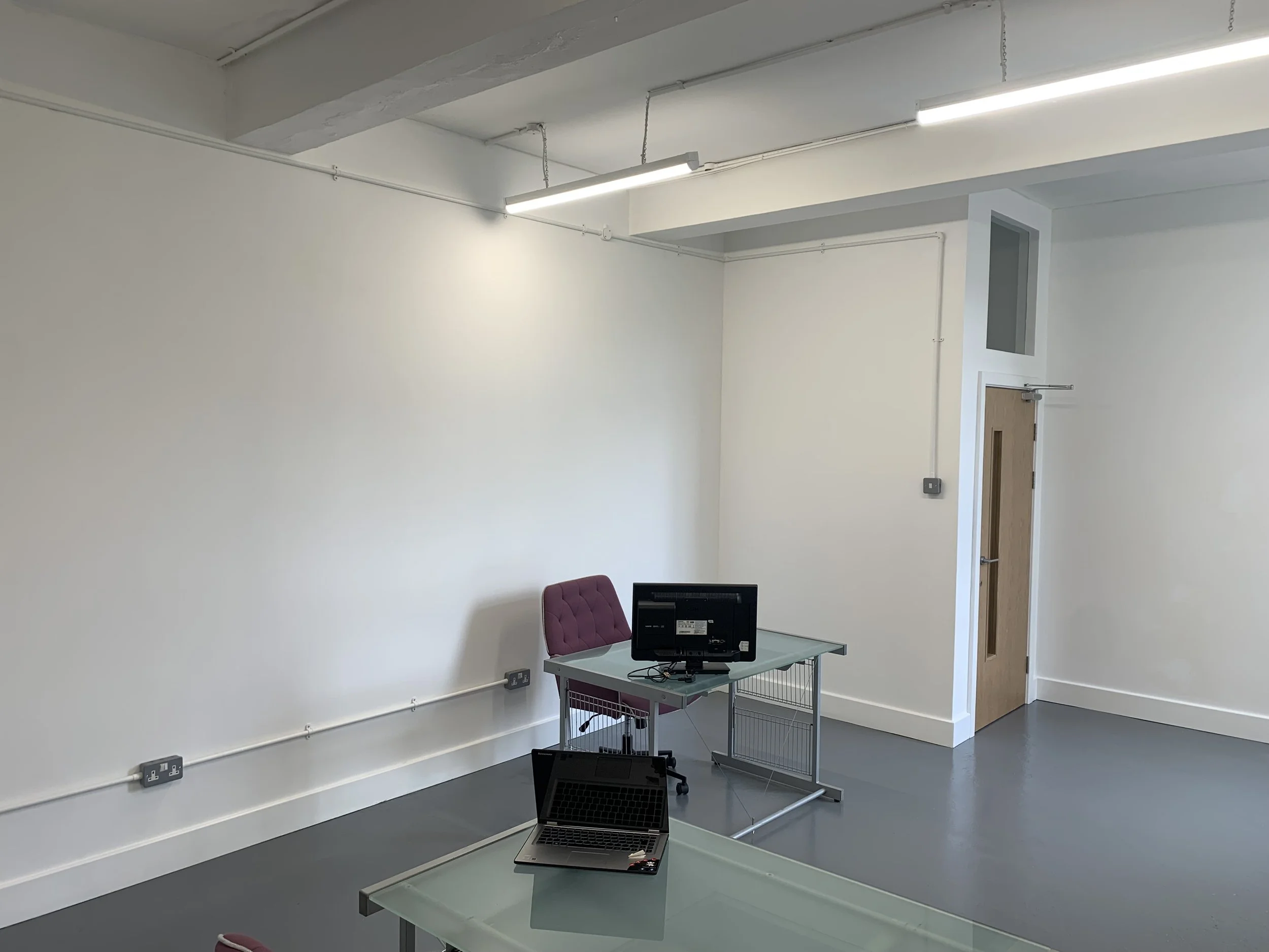 An empty office room with white walls, fluorescent lighting, a small desk with a laptop, a pink office chair, and a doorway with a wooden door.