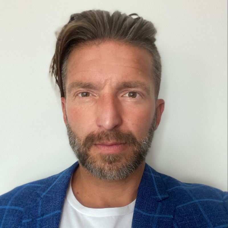 A man with styled brown hair, a beard, and blue eyes, wearing a blue blazer over a white shirt, standing in front of a white wall.