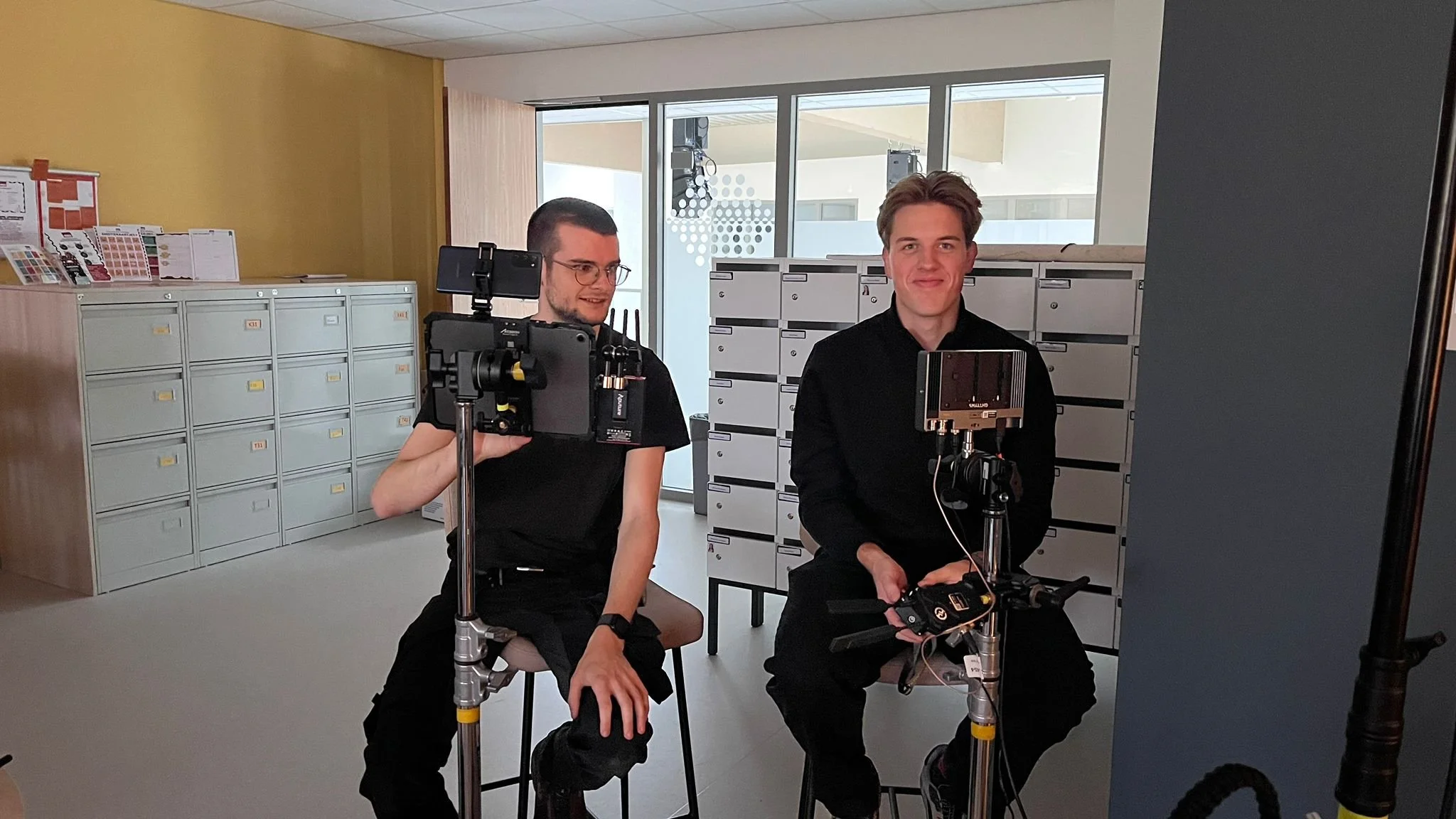 Twee jonge mannen filmen een filmpje met professionele camera's en stabilizers in een kantooromgeving met postvakjes en documenten op de achtergrond.
