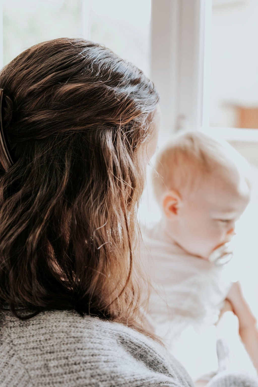 Renee, a baby and toddler sleep coach, with brown hair looking at her baby with blonde hair sitting by a window.