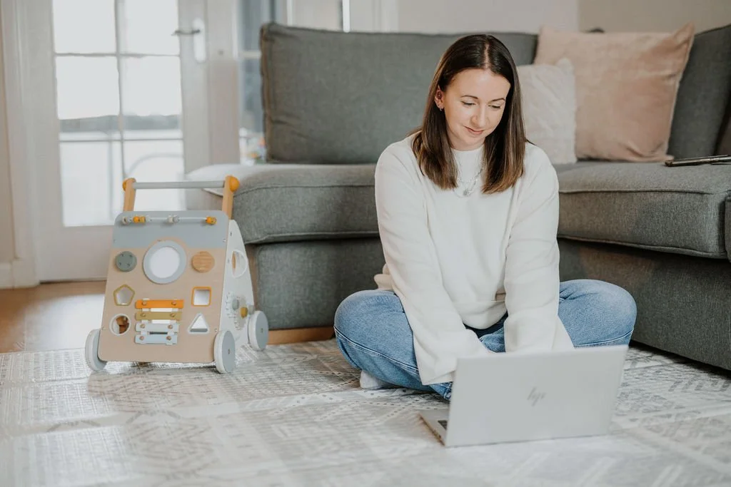 Renee, a baby and toddler sleep consultant, sitting on a patterned rug next to a modern activity cube toy, using a laptop, in a cozy living room with a gray sofa and pillows.