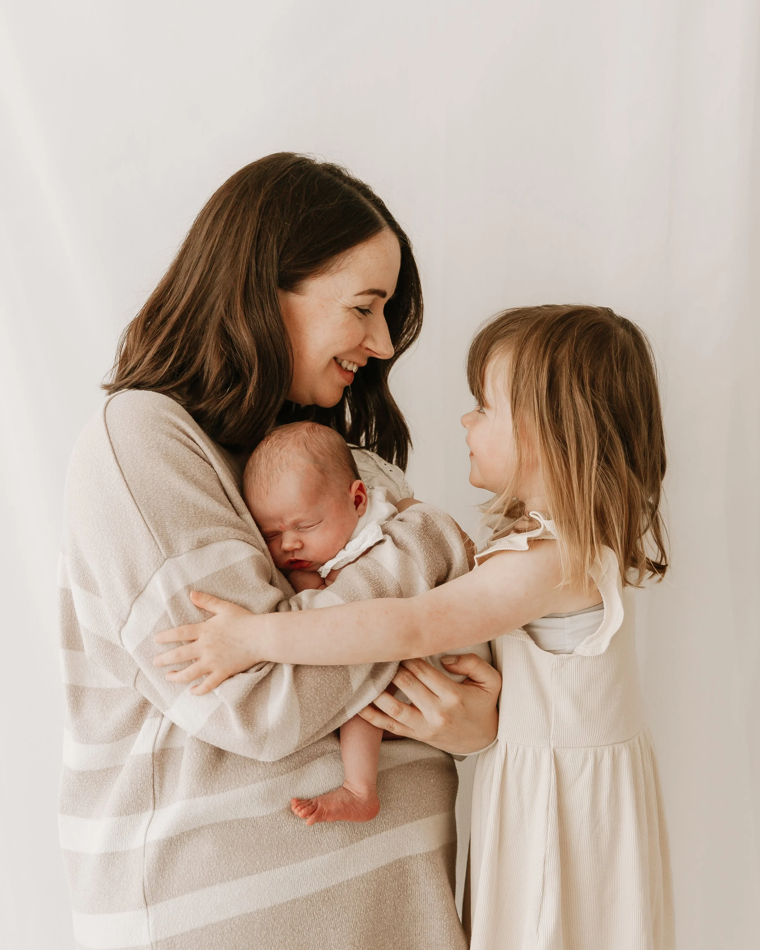 Renee, a baby and toddler sleep coach, with long brown hair holding her newborn baby, with her toddler with long blonde hair touching her baby's hand, both smiling and looking at each other, against a plain white background.