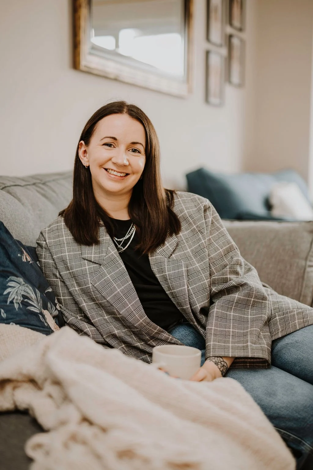 Renee, a baby and toddler sleep coach, with shoulder-length dark hair, wearing a plaid blazer and black shirt, sitting on a sofa in a cozy living room holding a mug.