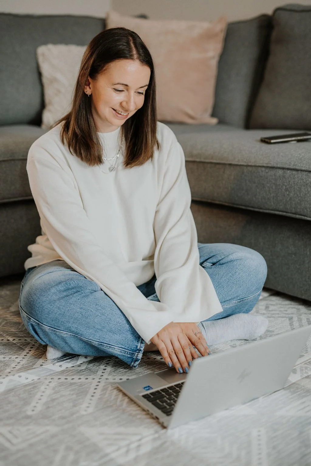 Renee, a baby and toddler sleep consultant sitting cross-legged on a patterned rug, smiling while looking at her laptop screen, in a cozy living room with a gray sofa and pillows in the background.