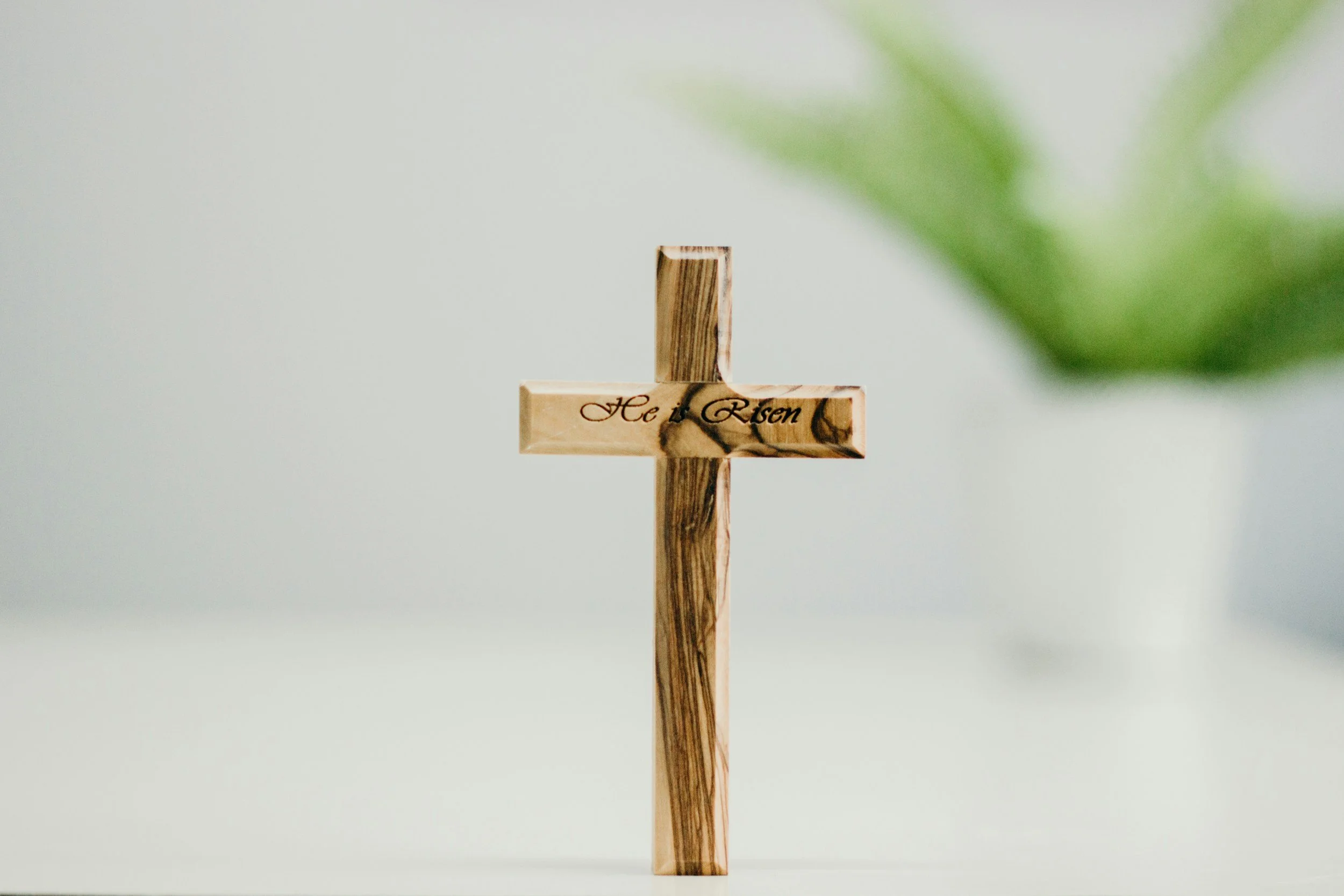 Wooden cross with the inscription 'He is Risen' engraved on the horizontal beam, with a blurred green leaf in the top right corner, set against a plain light background.