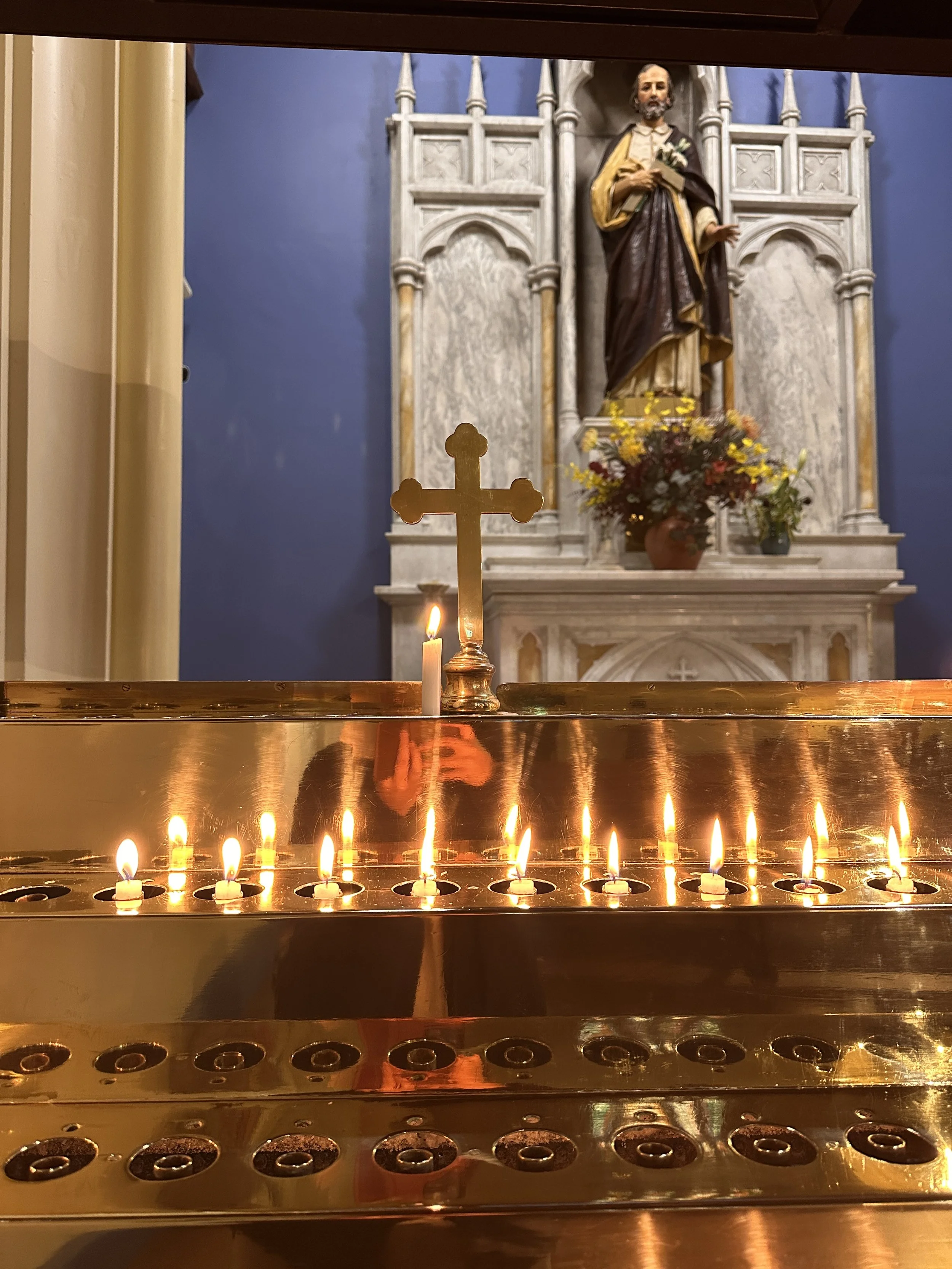 A church altar with multiple lit candles, a gold cross in the foreground, a statue of a saint holding flowers and a book in the background, and flowers in a vase on the altar.
